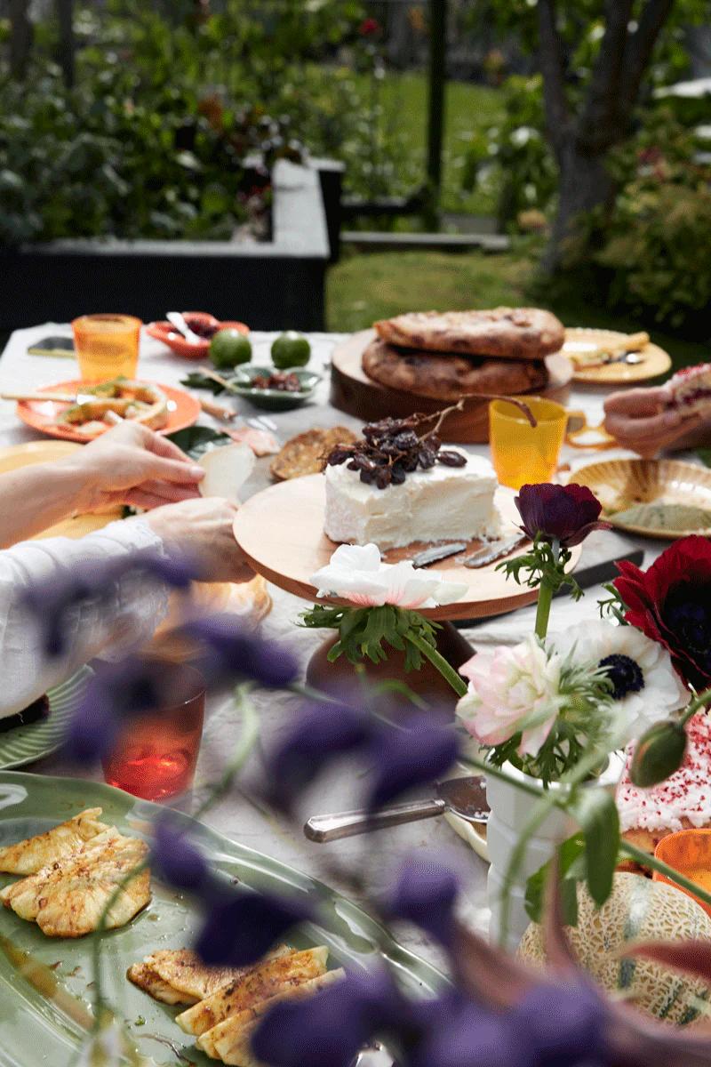 An outdoor table features caramelised onion cheese, flatbreads, fruit, and drinks. Greenery and flowers create a fresh al fresco setting.