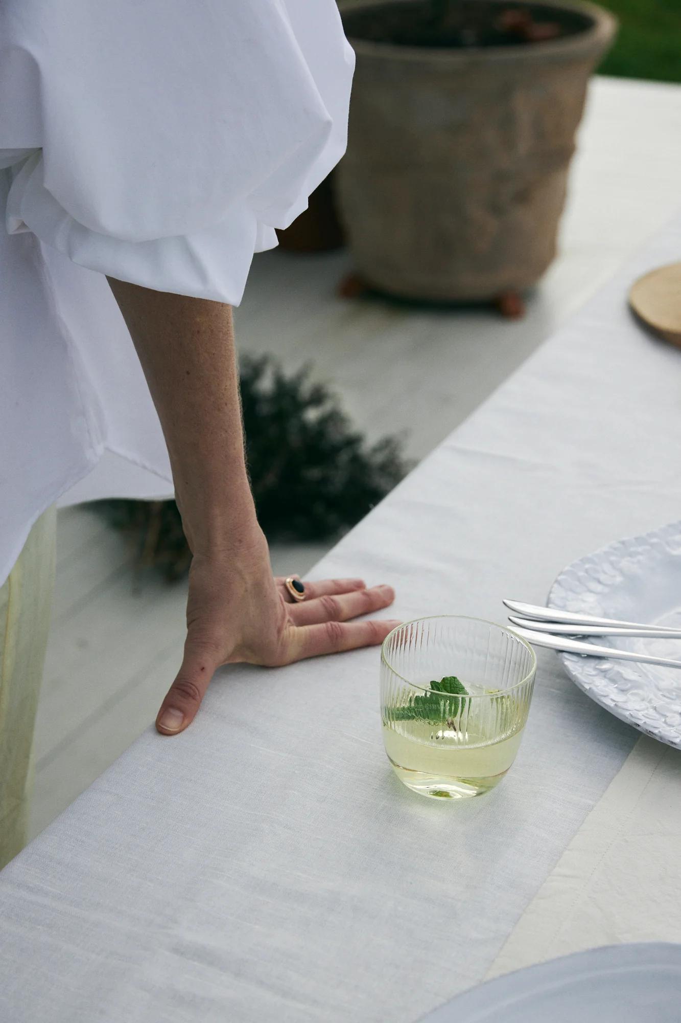 Person in white blouse rests hand on table. Partially filled green drink, white plate, and cutlery nearby.