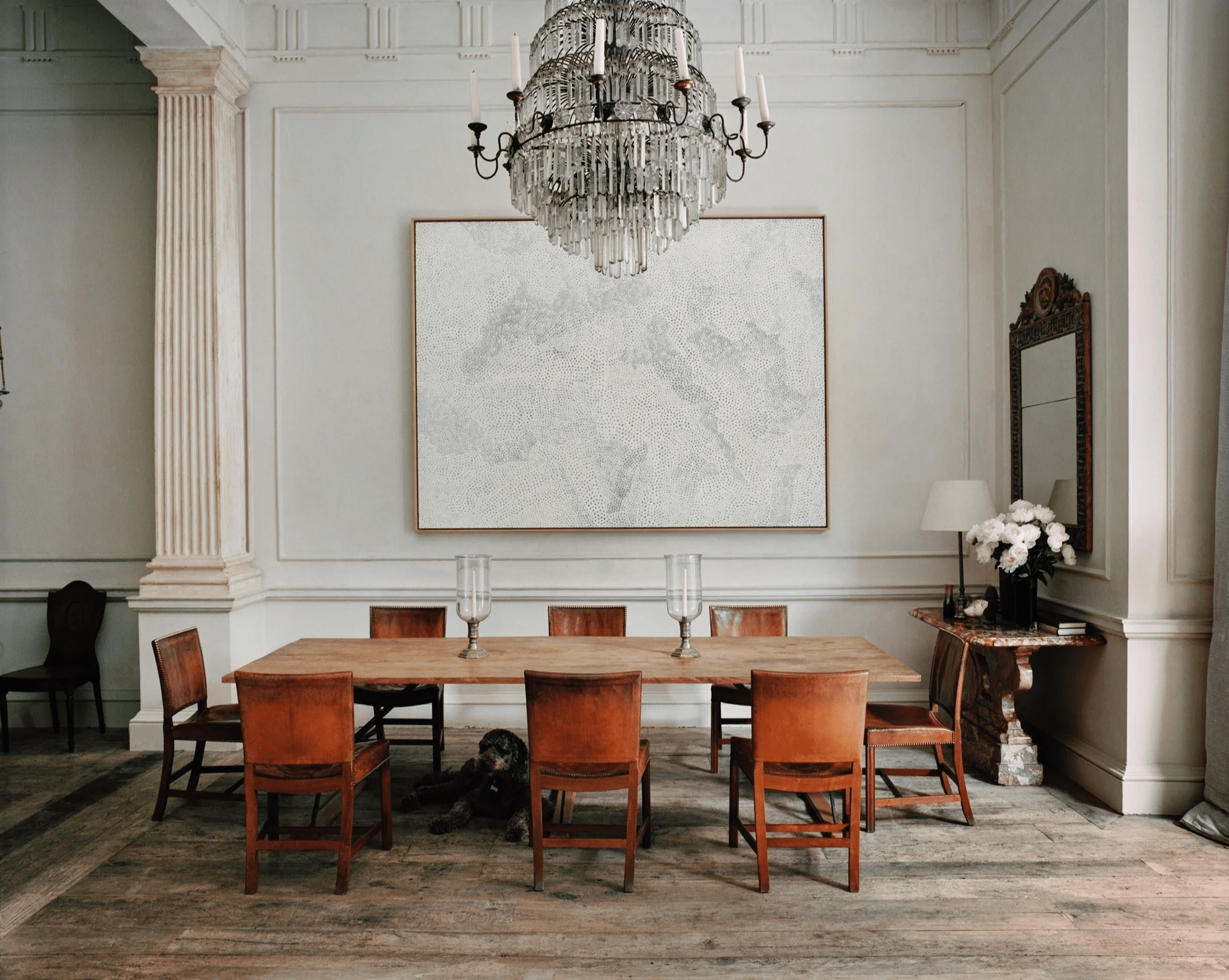 Wooden dining table with brown chairs. Crystal chandelier, abstract painting. Dog near table, flowers on side.