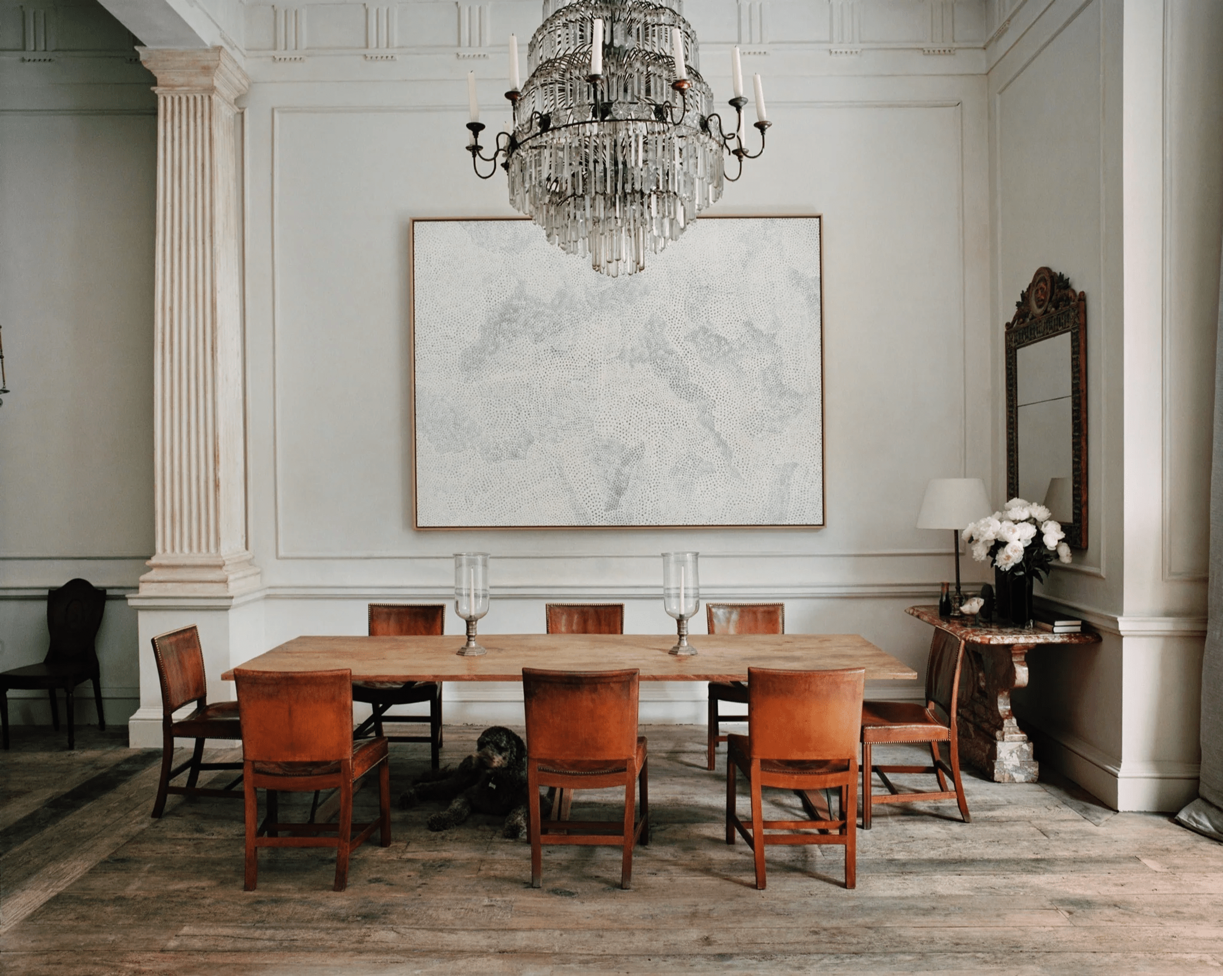 Wooden dining table with brown chairs. Crystal chandelier, abstract painting. Dog near table, flowers on side.