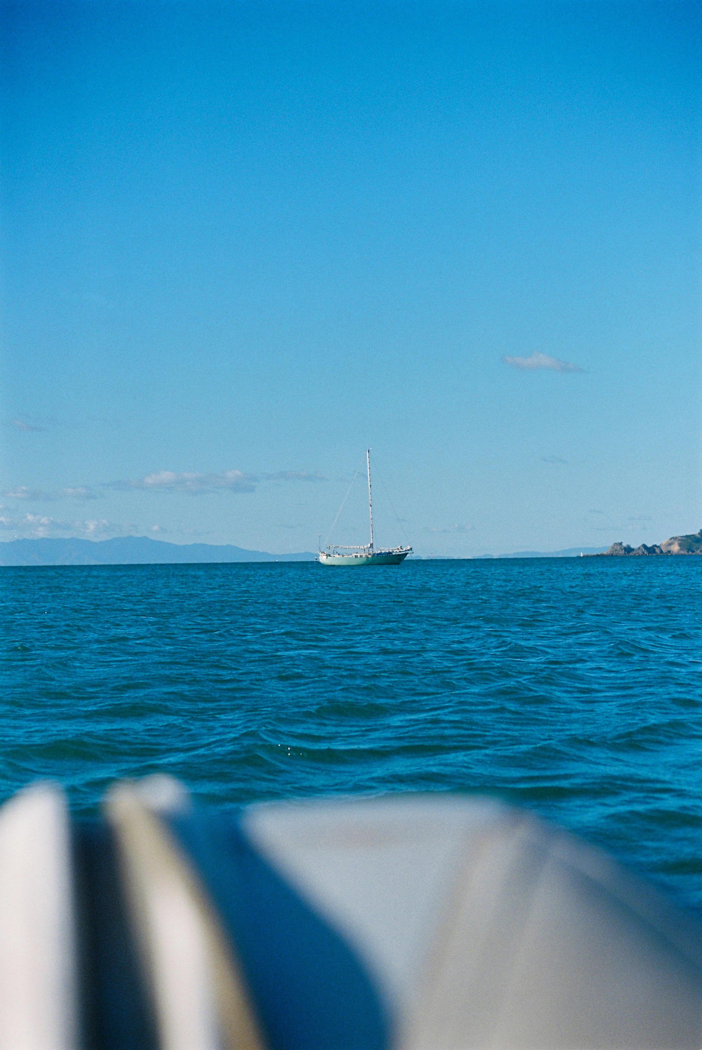 Distant sailboat on calm waters. Blurred foreground, land with hills on horizon.