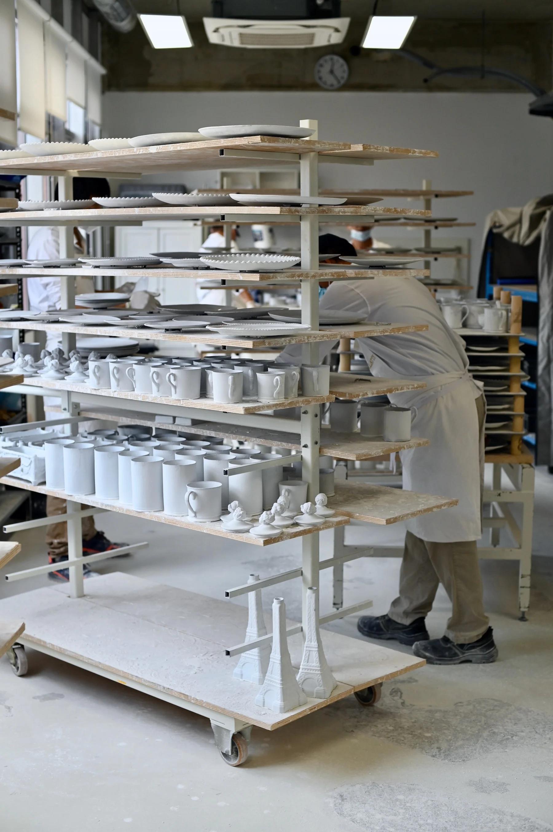 Person in lab coat arranging white ceramic mugs and Eiffel Tower replicas on multi-tiered rack. White dust on floor, other workers in background.