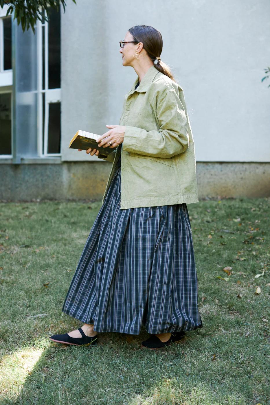 Person with glasses in green top and plaid skirt. Holding book, standing on grass near building with large windows.