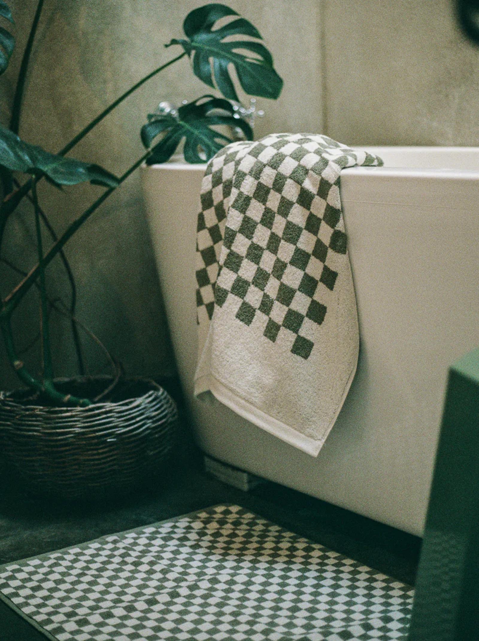 A green and white checkered towel draped over a white bathtub, with a matching bathmat and a potted plant beside it against a neutral stone wall.