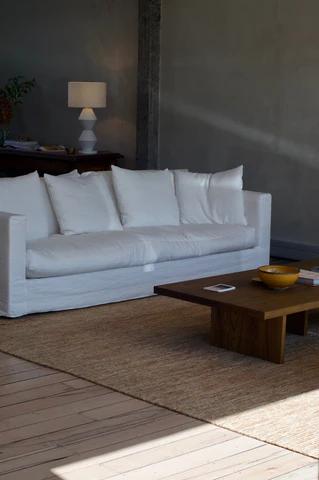 Minimalist living room with white sofa, wooden table, and textured rug. Sideboard with lamp and plant against grey wall.