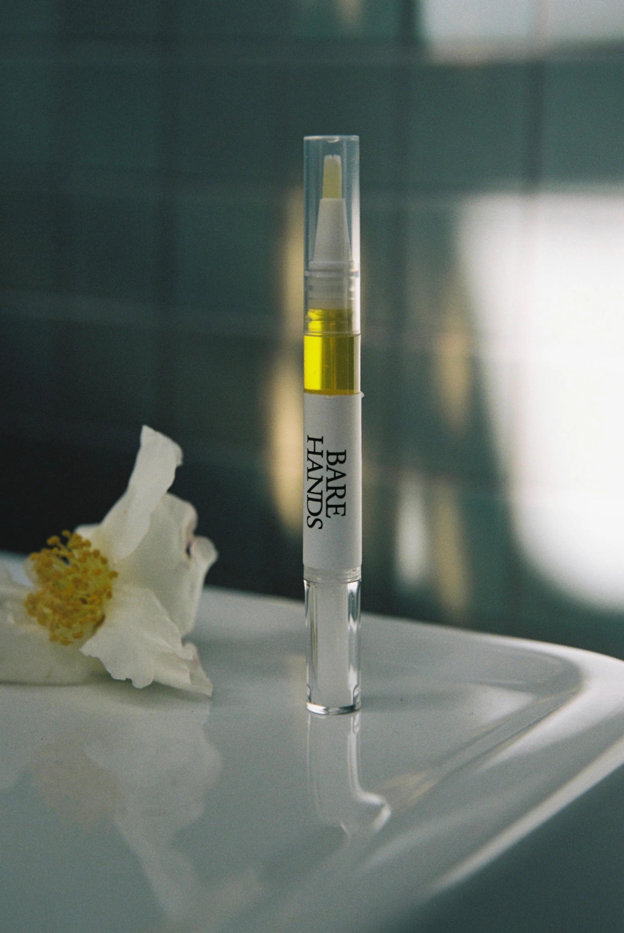 A sleek, clear tube of Bare Hands cuticle serum stands on a glossy bathroom vanity. The label is visible, and a white flower lies next to the tube, with a blurred tiled background.