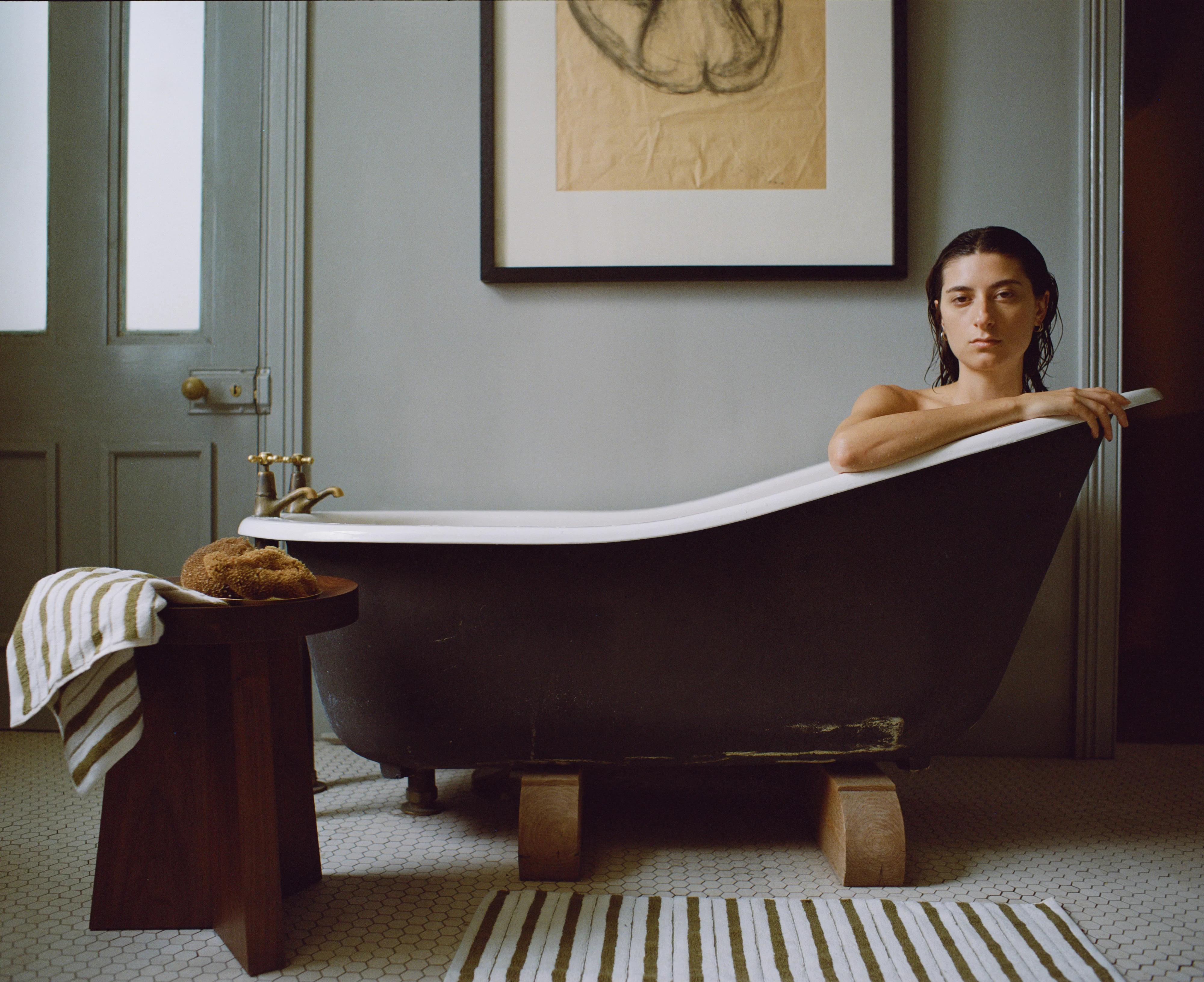 Person in vintage black clawfoot tub. Hexagonal tile floor, striped towel on stool, striped rug, framed art.