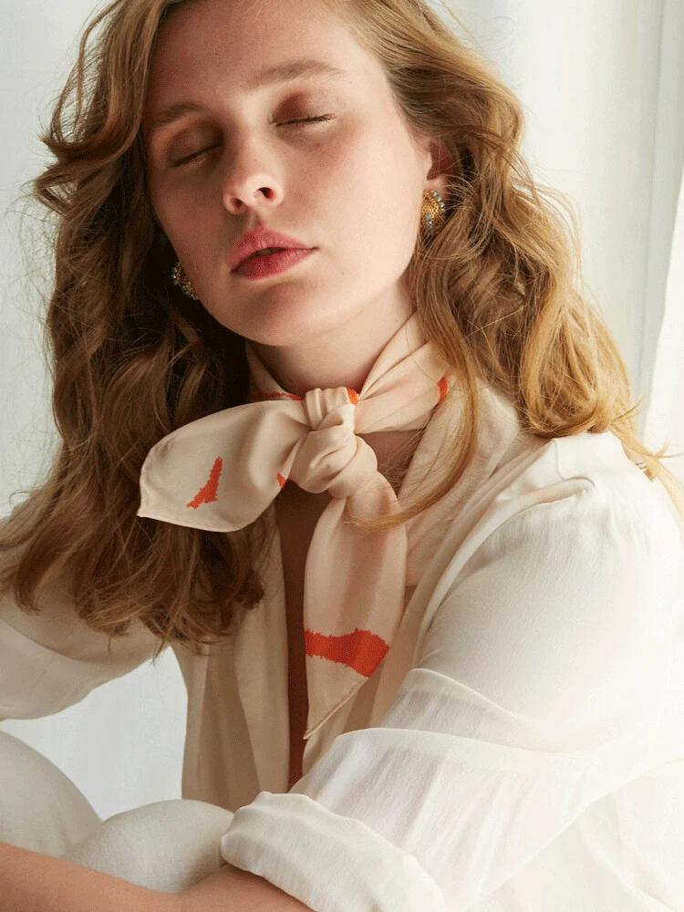 Woman with wavy hair sits with eyes closed, wearing cream blouse and orange Walker & Bing scarf. White curtains in background create serene atmosphere.