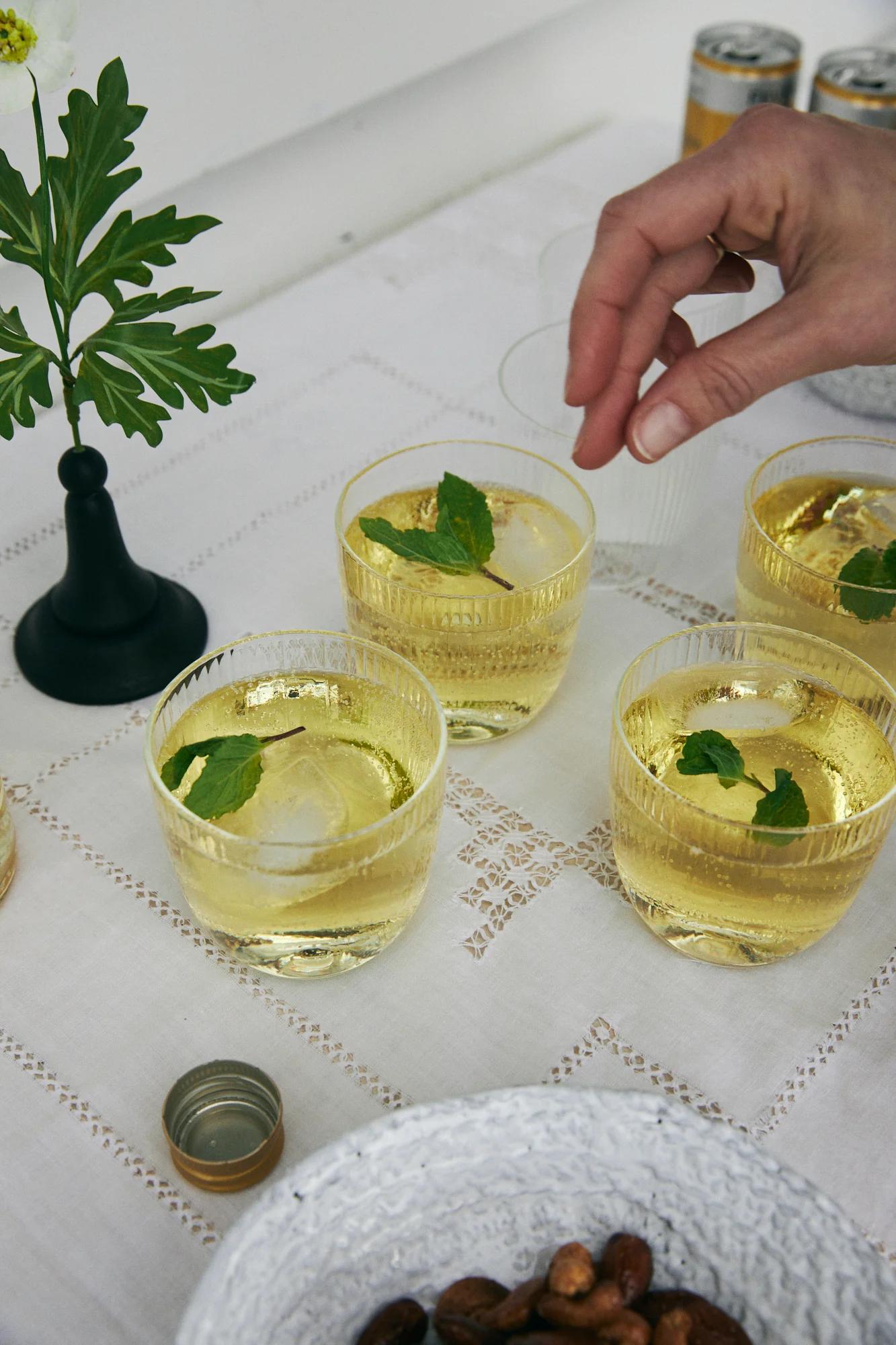 Person garnishing drink with mint leaf. Three glasses with yellow liquid and ice. Flower vase and nut bowl on tablecloth.