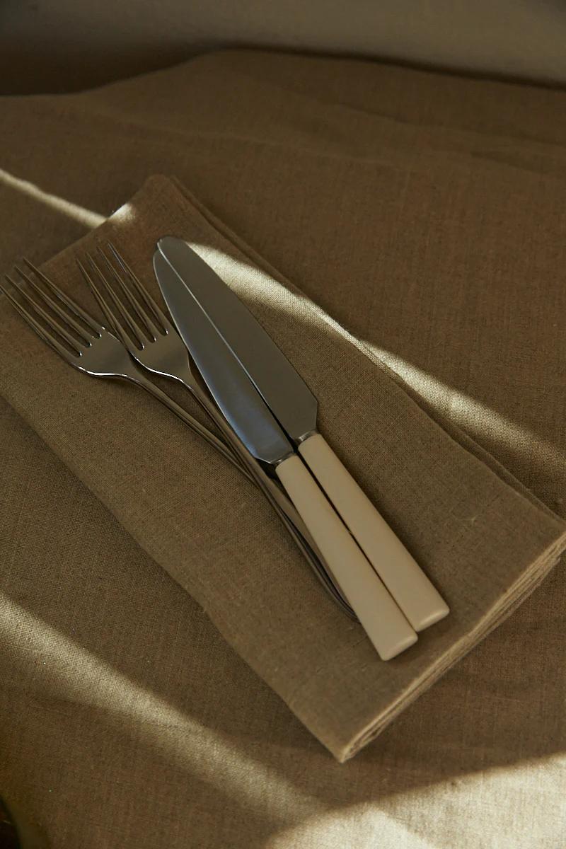 Beige-handled forks and knife on folded brown Libeco napkin. Beige surface with soft sunlight shadows.