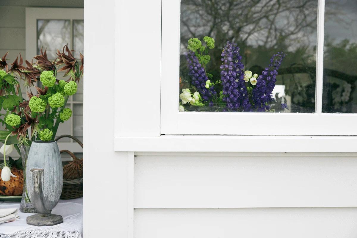 White structure reflects garden. Window displays purple and green flowers. Outside, vase with green flowers on table evokes serene holiday setting.