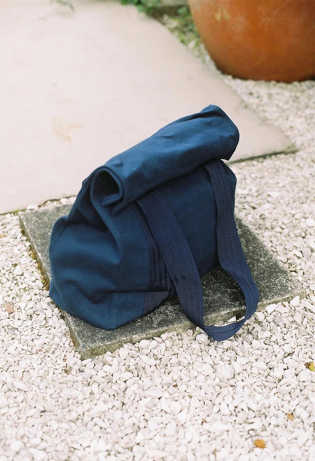 Blue canvas bag with dark straps on concrete slab amid gravel. Folded top creates casual look. Large brown planter visible in corner.