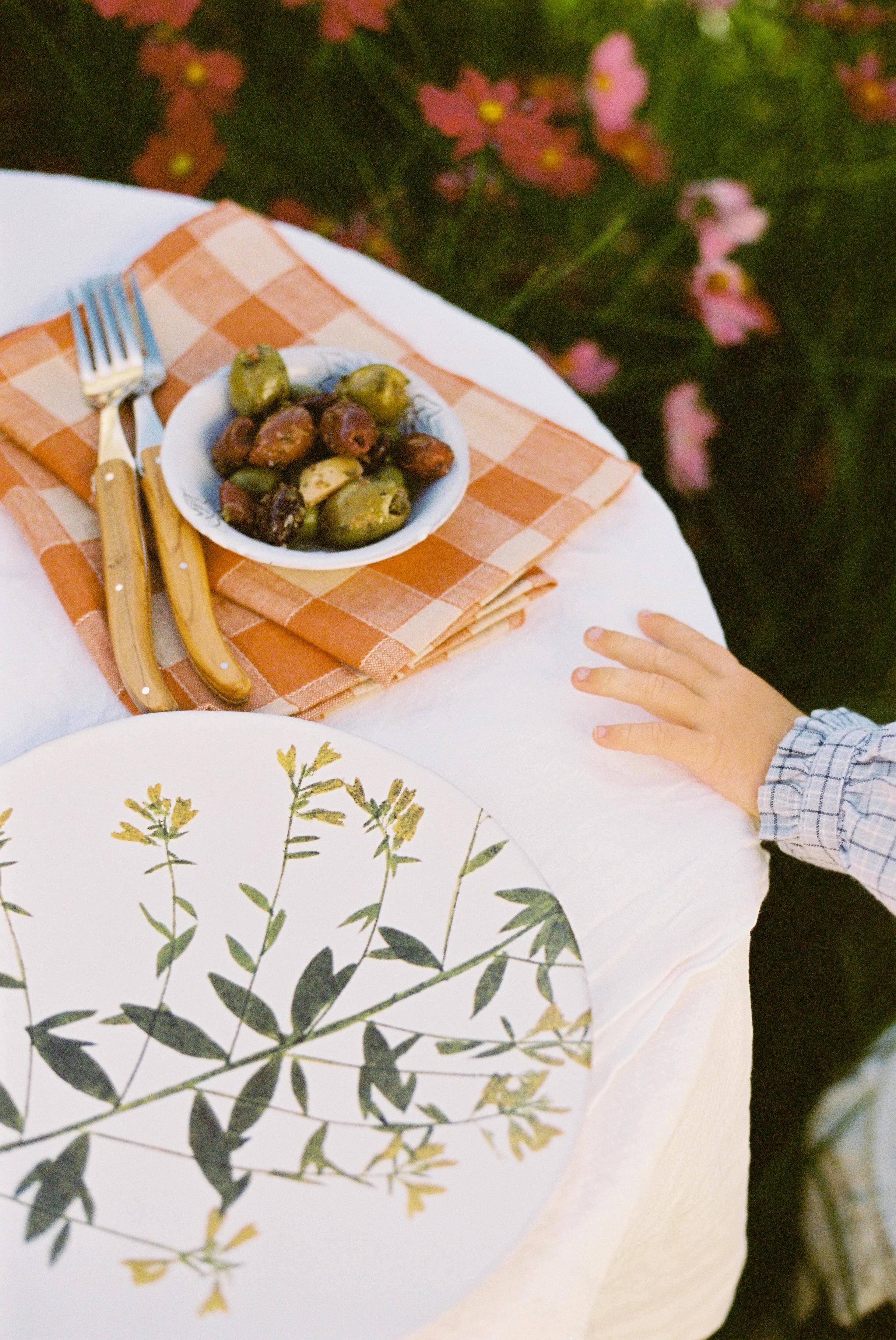 Child's hand reaching for a plate with green frittata. Table set with orange napkin, cutlery, and olives. Pink flowers and greenery in background capture spring essence.