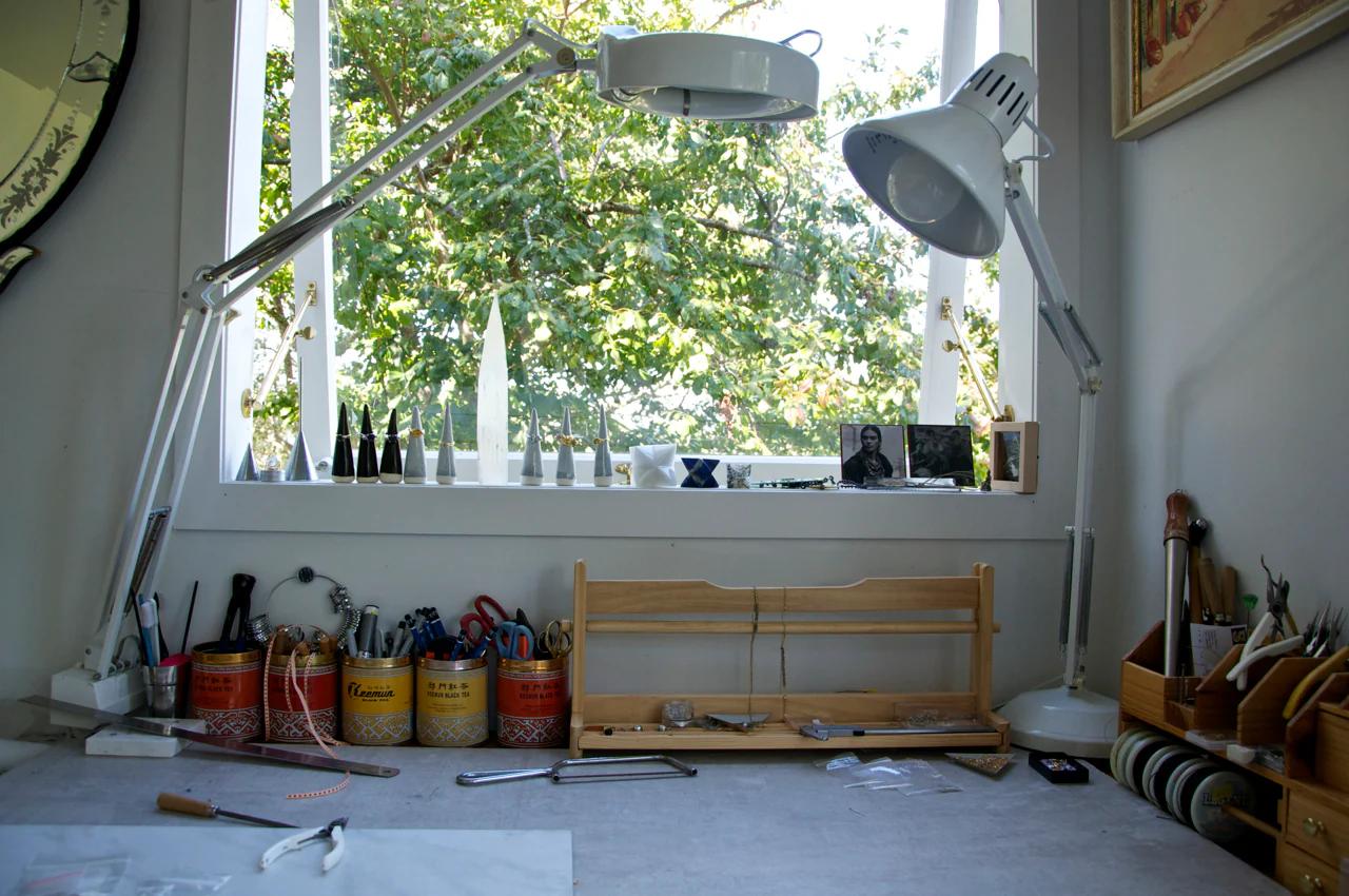 Organised home studio desk with tools, lamps, and supplies. Window shows green foliage. Shelves hold containers and small items. Partially assembled project on desk.