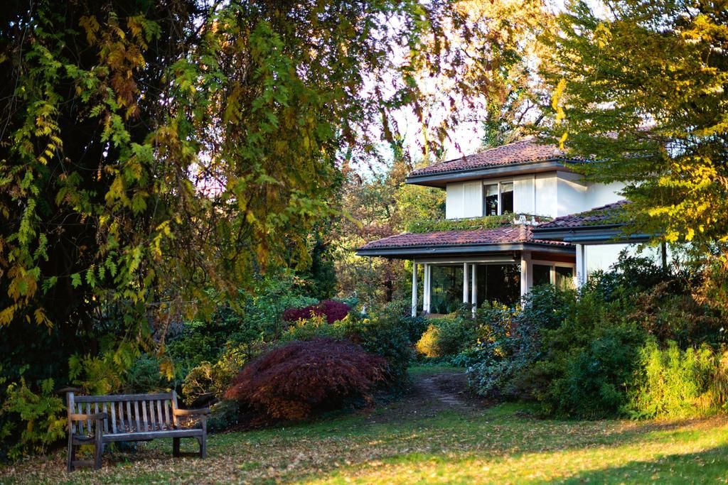 Two-story house with red roof among greenery. Wooden bench under tree. Pathway to porch surrounded by vibrant garden.