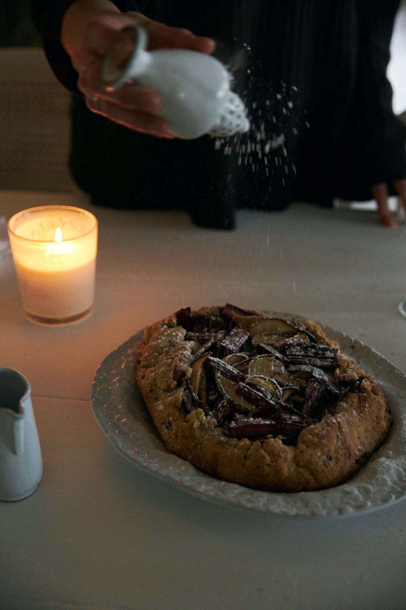 Person sprinkling sugar on tart. Glowing candle creating cozy ambiance. Small white pitcher nearby.