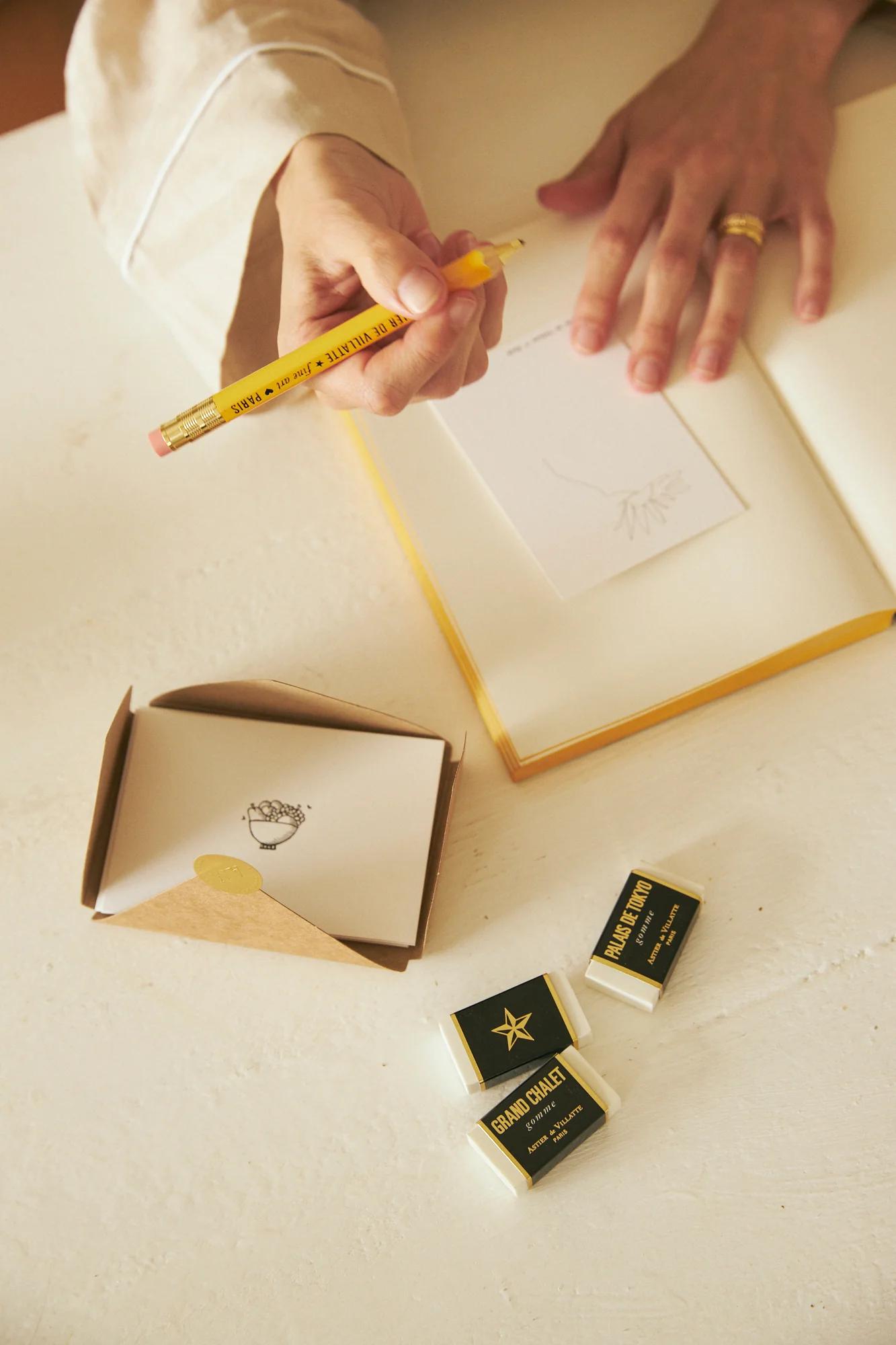 Person drawing in notebook. Boxes labelled "GRAND COAST" and open package with floral stamp nearby.