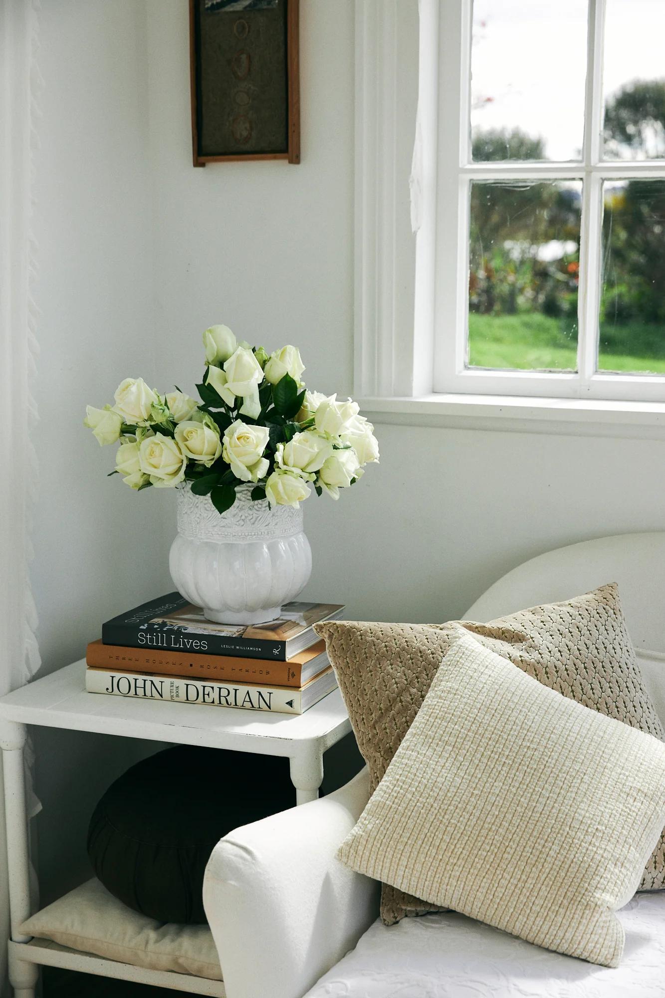 White corner with textured pillows on chair. Side table with roses, books, and Astier de Villatte ceramics. Window shows garden view.