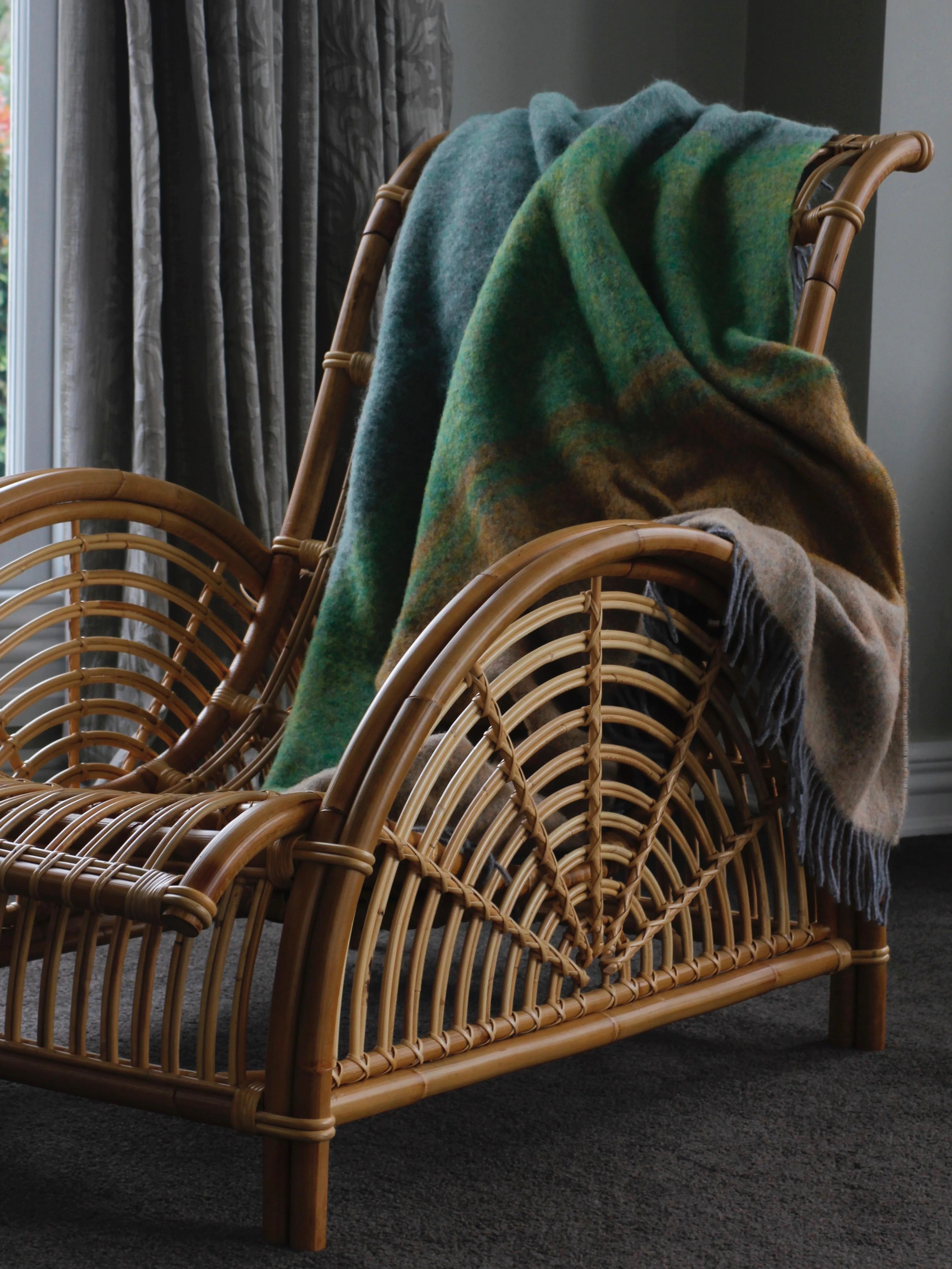 A wicker chair with a green and brown wool blanket draped over it, placed near a window with grey curtains on a dark grey carpeted floor.