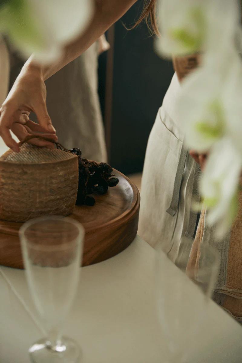 Person slicing bread on wooden board. Empty champagne flute nearby, white flowers in foreground.