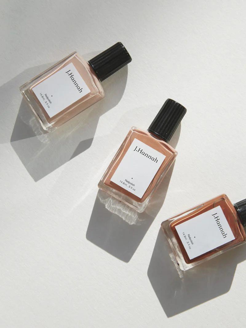 Three bottles of J. Hannah nail polish lie on a light surface, casting soft shadows. The bottles feature black caps and white labels, with neutral beige tones inside.