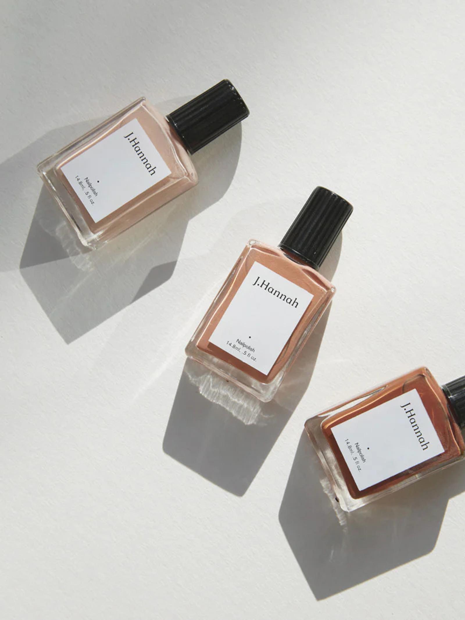 Three bottles of J. Hannah nail polish lie on a light surface, casting soft shadows. The bottles feature black caps and white labels, with neutral beige tones inside.