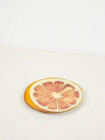 Ceramic plate mimics halved orange with detailed rind and fruit sections. Placed on white surface, it evokes sunny afternoons and refreshing drinks.