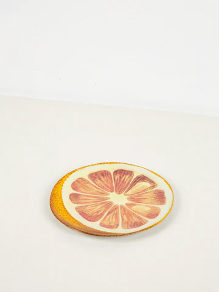 Ceramic plate mimics halved orange with detailed rind and fruit sections. Placed on white surface, it evokes sunny afternoons and refreshing drinks.