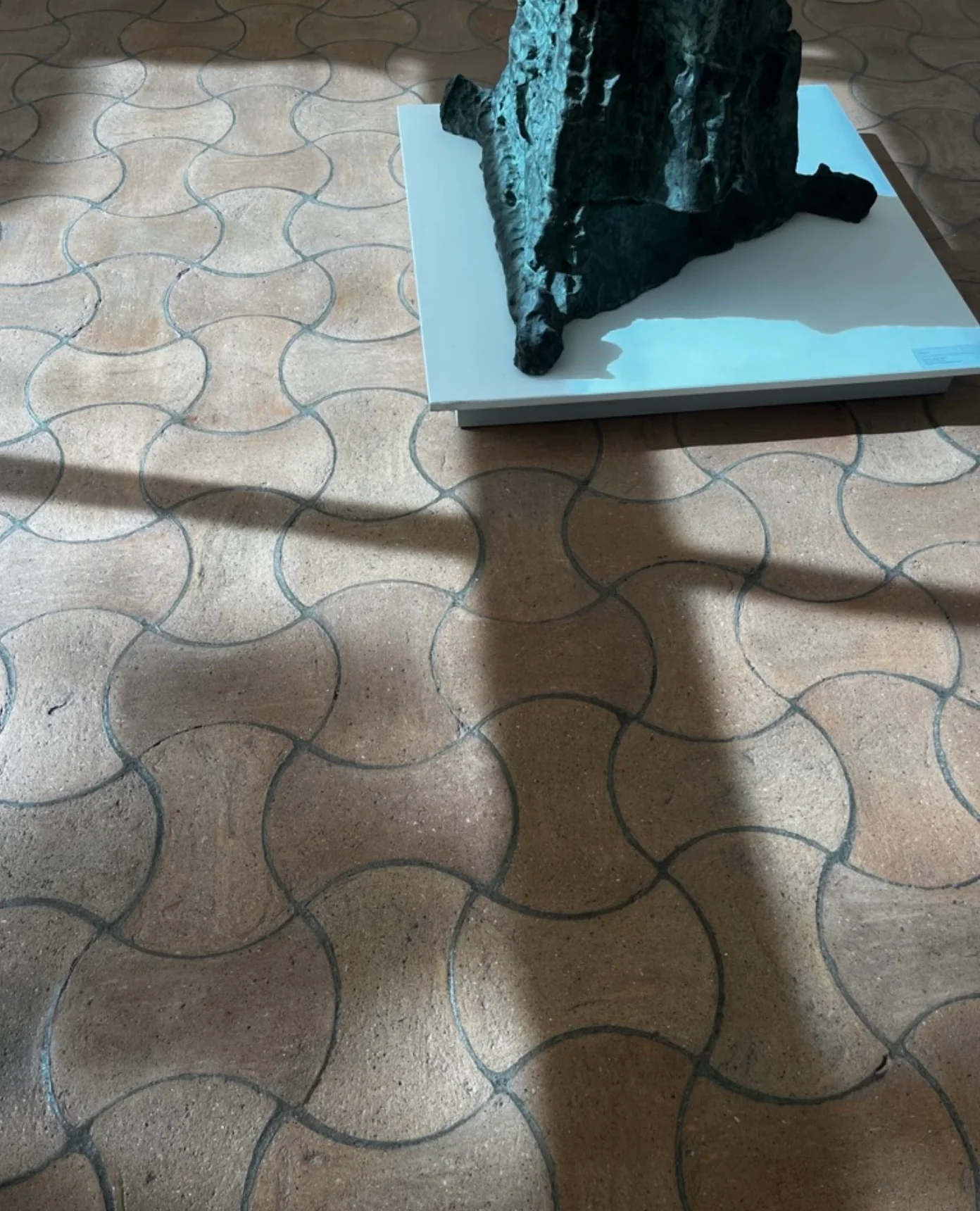 Window shadow on hexagonal terracotta tiles. Partial view of sculpture base. Natural light illumination.