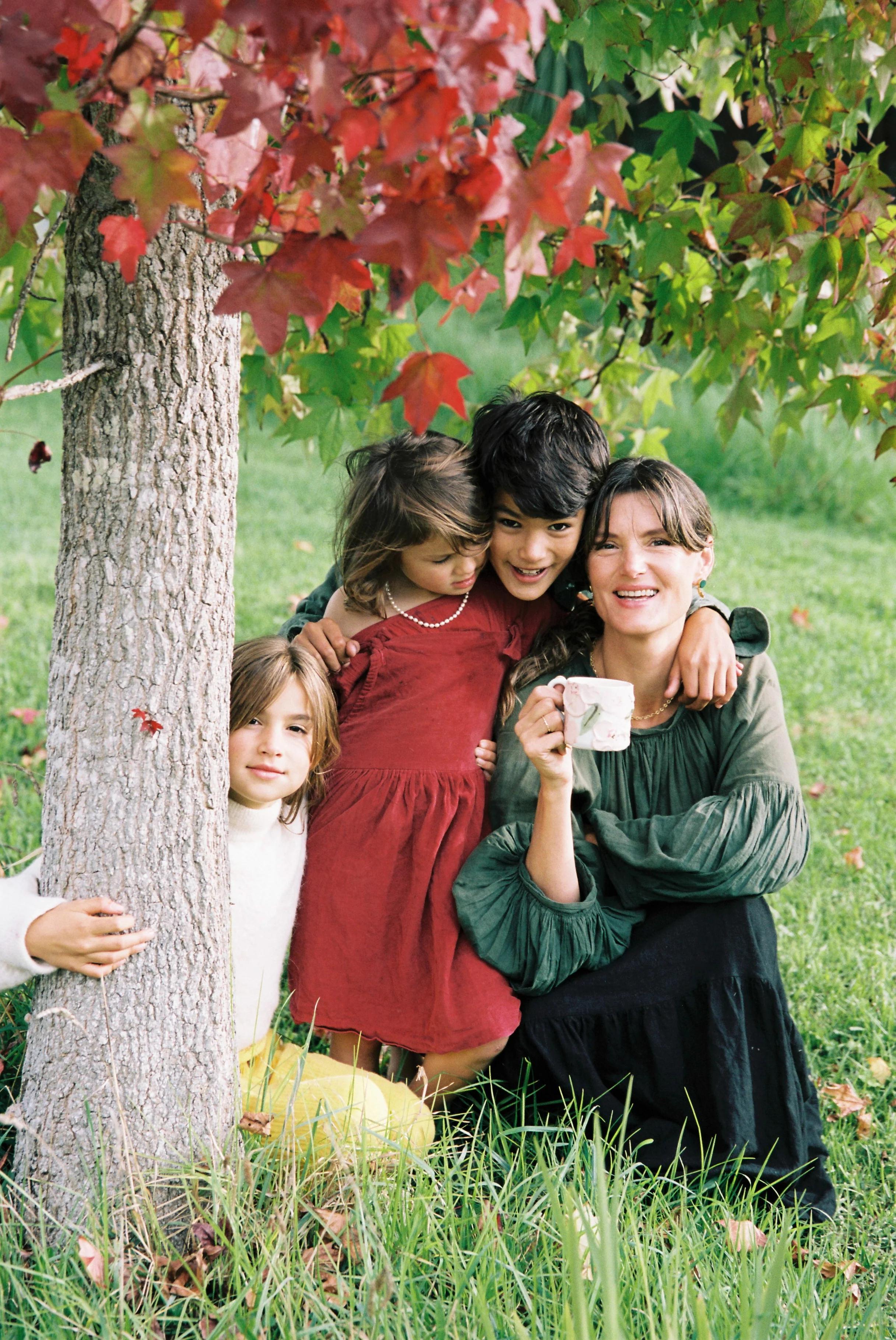 A mother and three children pose by a tree in a grassy field. She holds a floral mug. One girl wraps her arm around the tree, while the boy hugs the other girl and his mother.