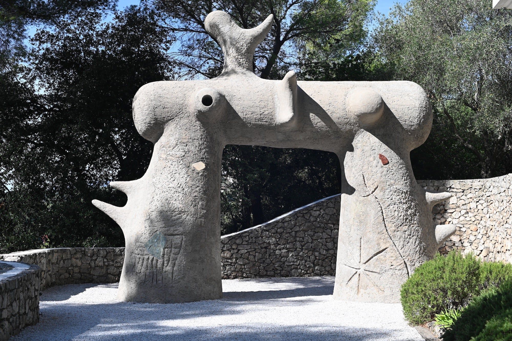 Large stone sculpture with arch shape and painted symbols. Sunny garden with trees and stone wall.