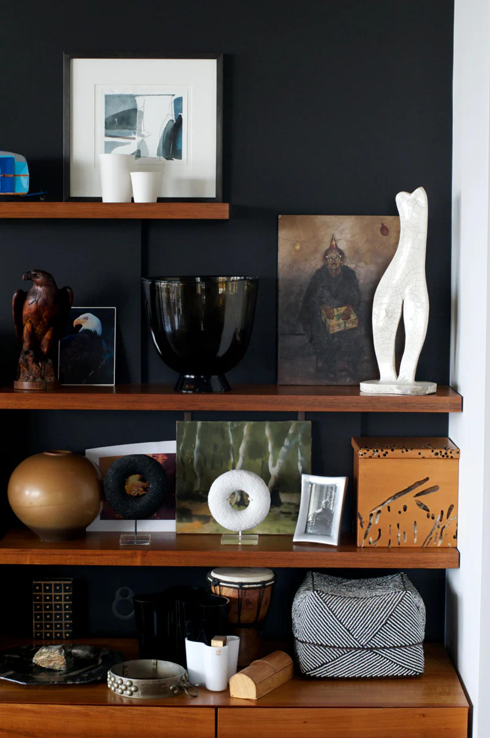 Wooden shelving unit displays eclectic decor against dark wall. Items include sculptures, artwork, bowls, and decorative objects in artistic arrangement.