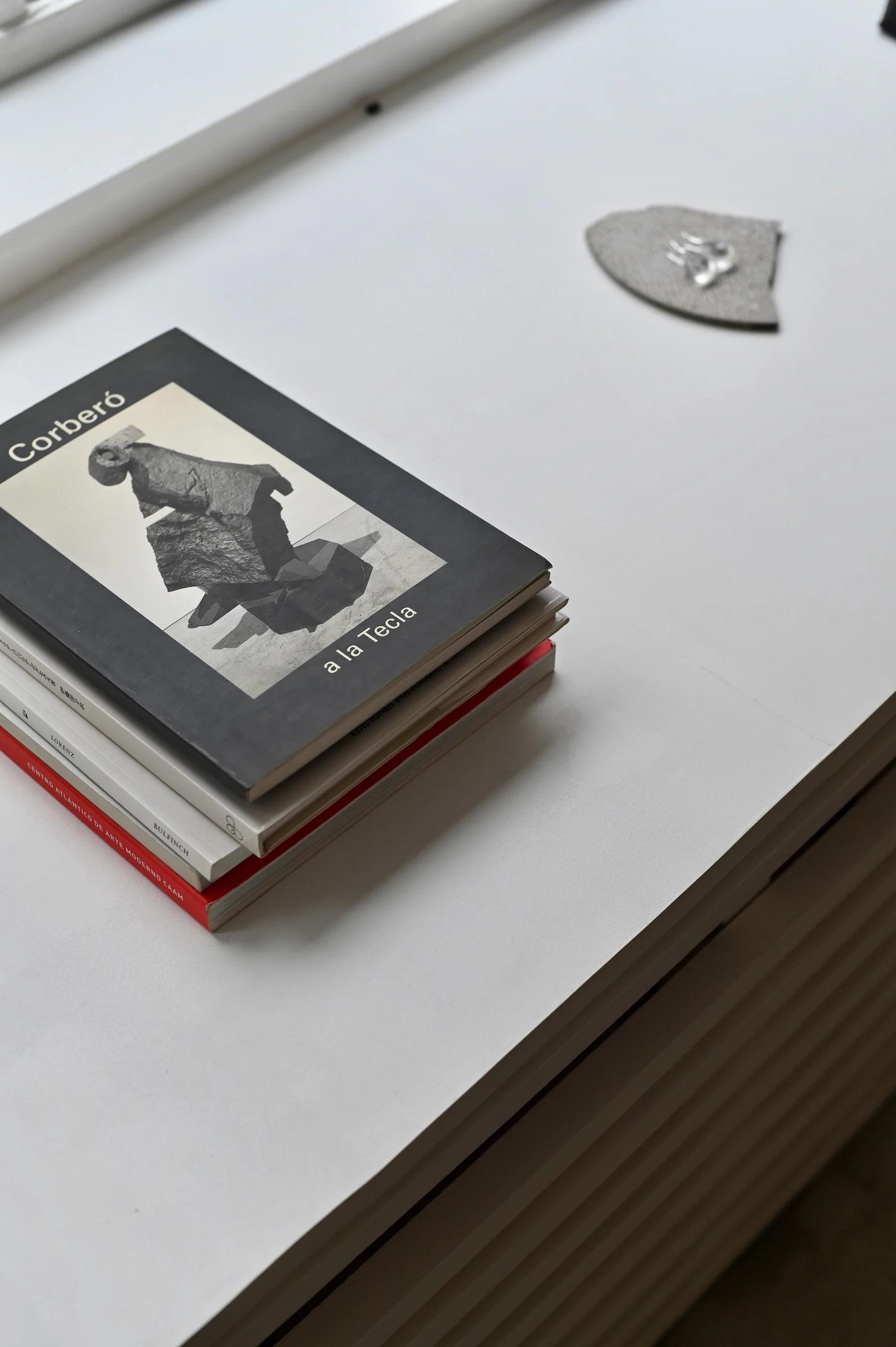 Stack of books on white table. Top book titled "Corberó" with black and white sculpture image. Small grey object in background.