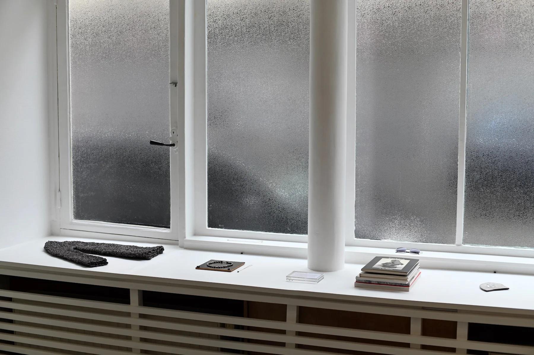 Frosted glass window behind white windowsill. Grey knitted gloves, trinket, books, and coaster on sill. White pillar to right.