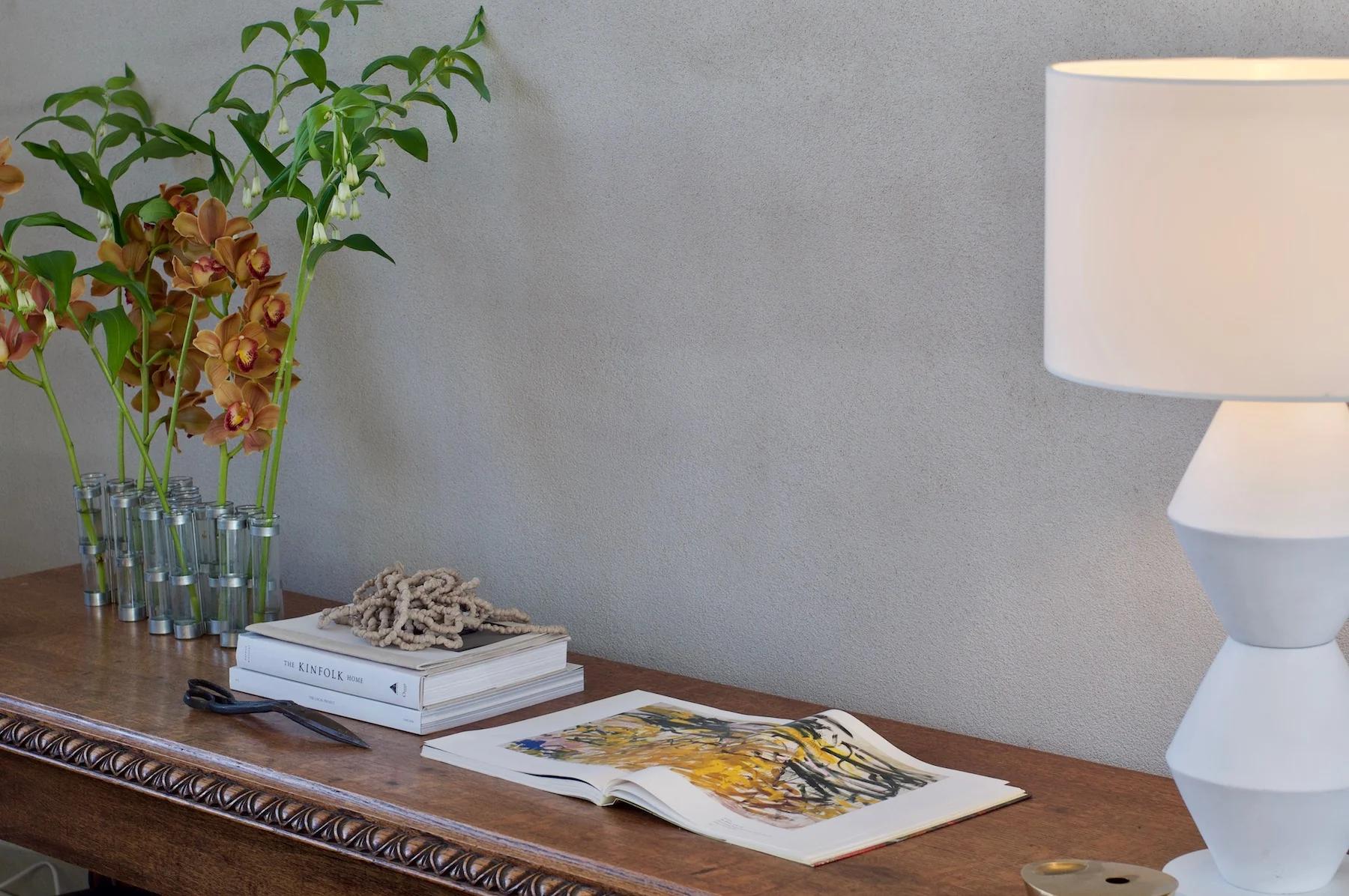 Wooden table with lamp, book, scissors, and rope. Vase of orange orchids on left, against plain white wall.