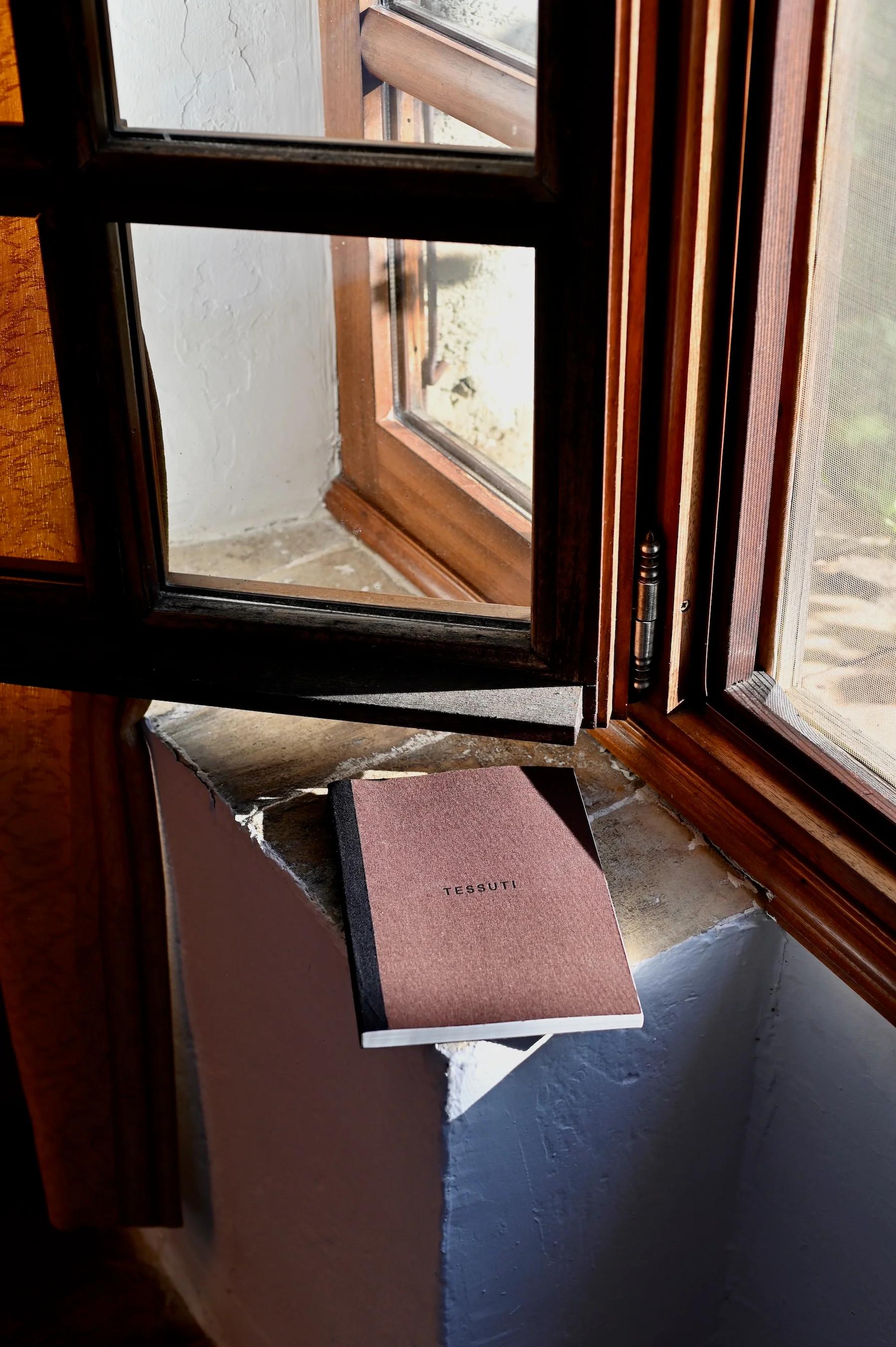 Brown "TESSUTI" notebook on stone windowsill. Sunlight through open wooden window.