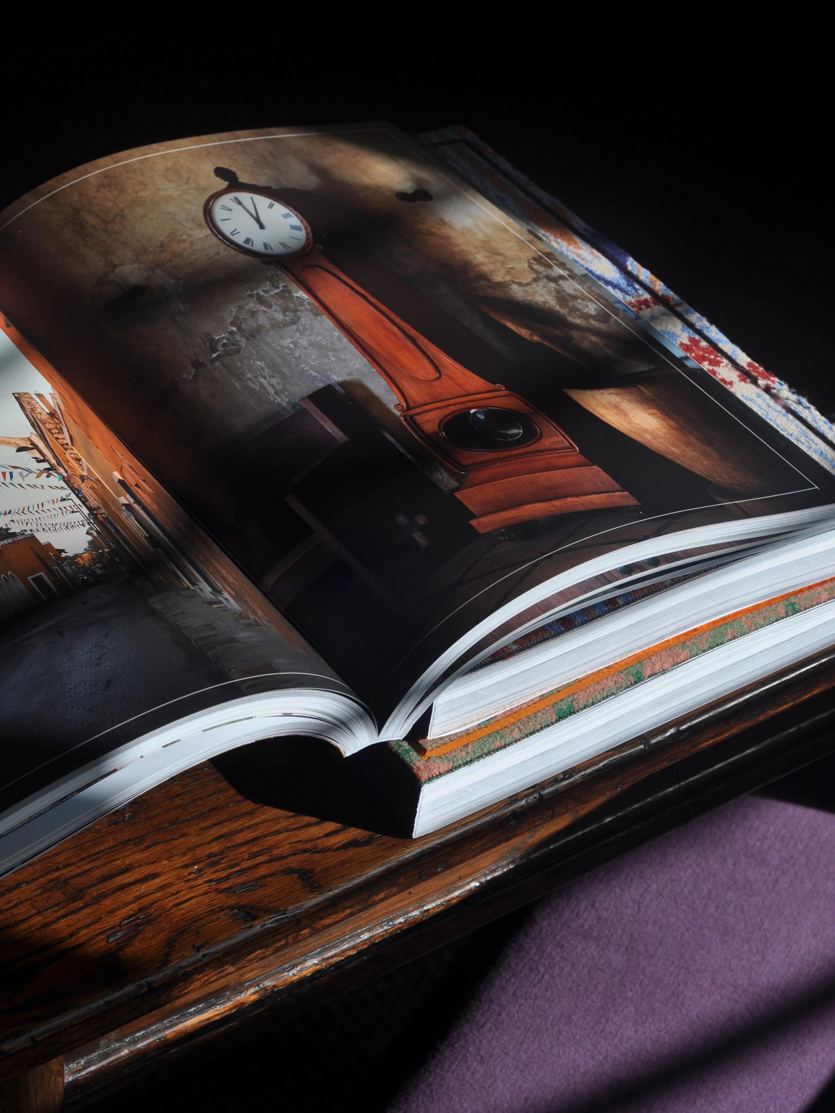 Open book showing wooden clock photo. Patterned cloth beneath, purple fabric in foreground. Wooden table, beam of light.