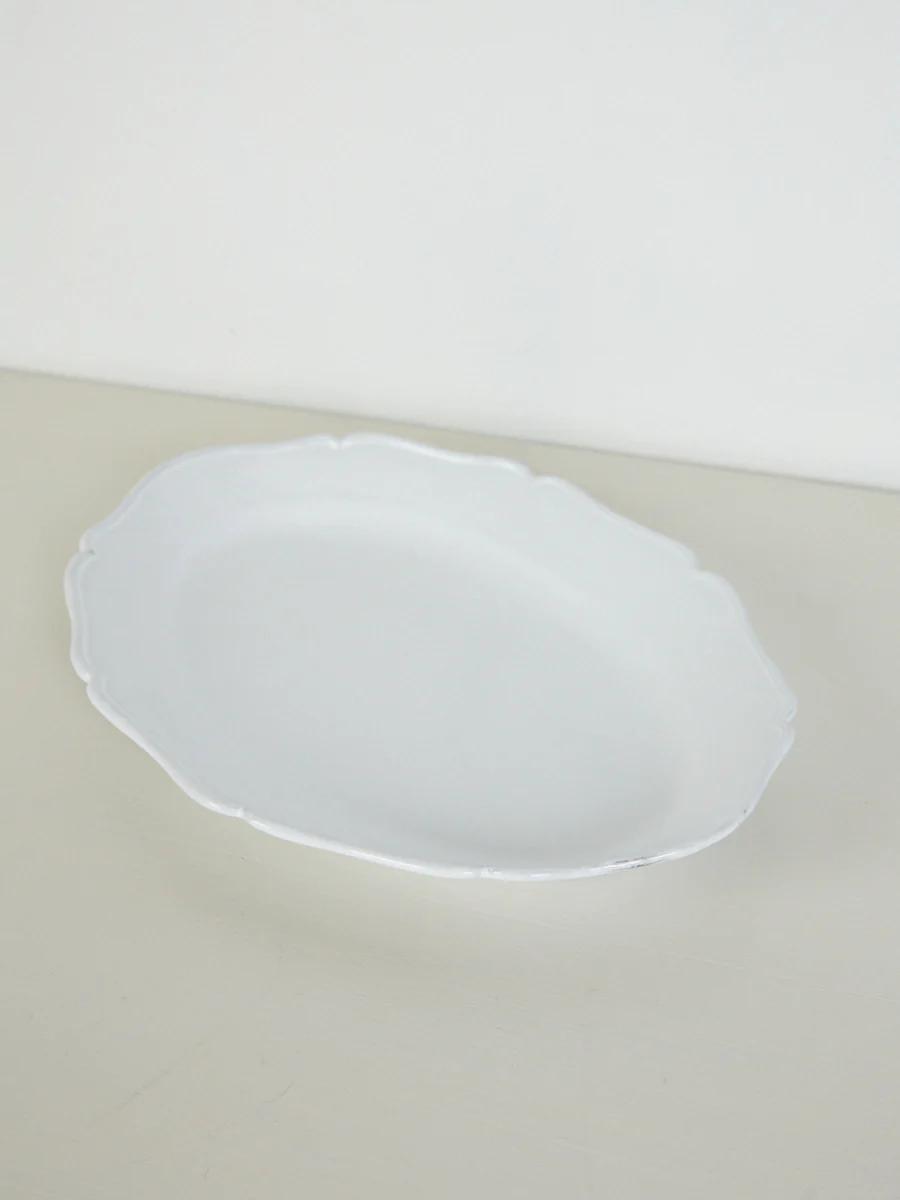 Empty white oval porcelain plate with scalloped edges on light beige surface. White background.