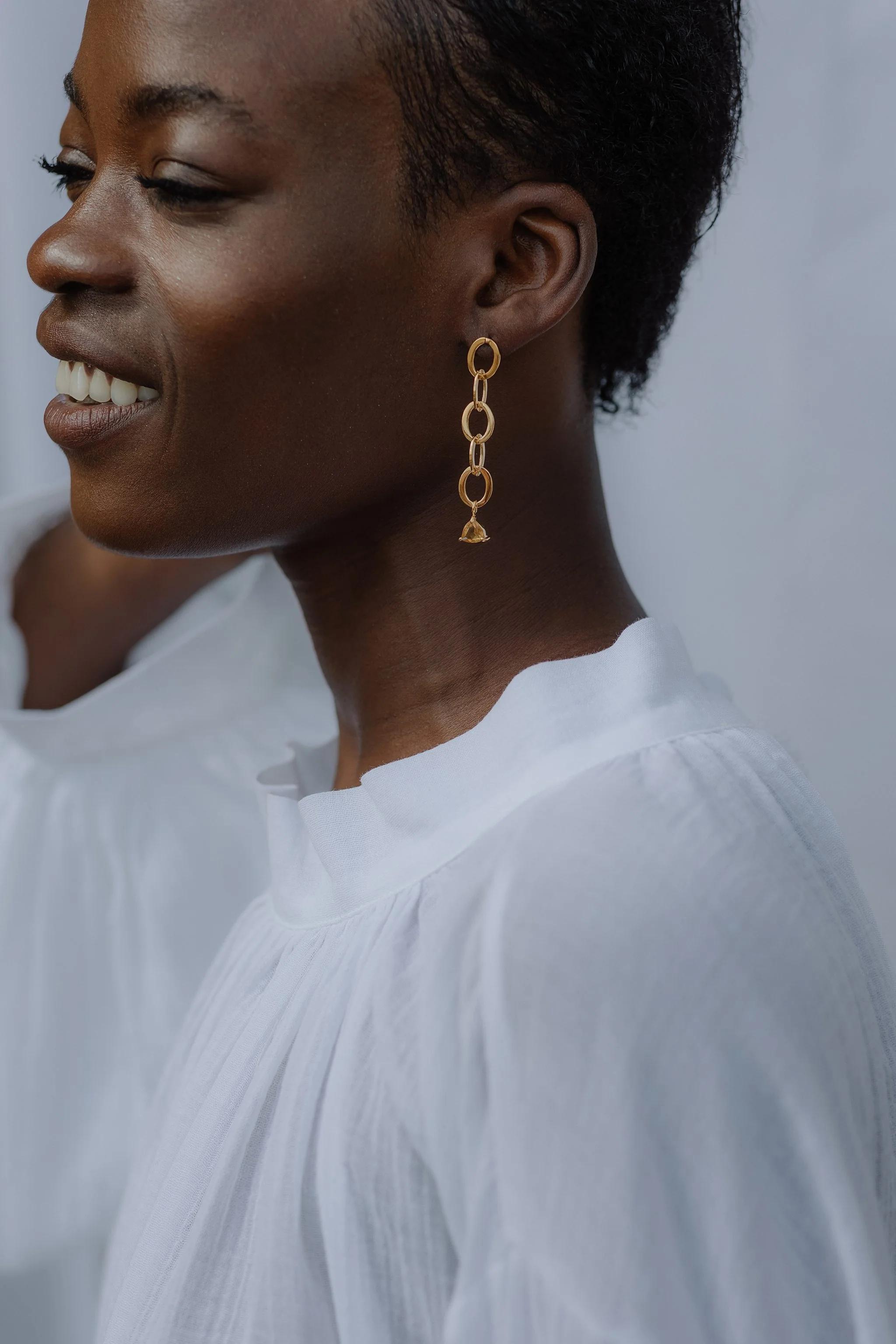 Person in white shirt smiling, wearing long dangling gold earring. Short hair, light background.