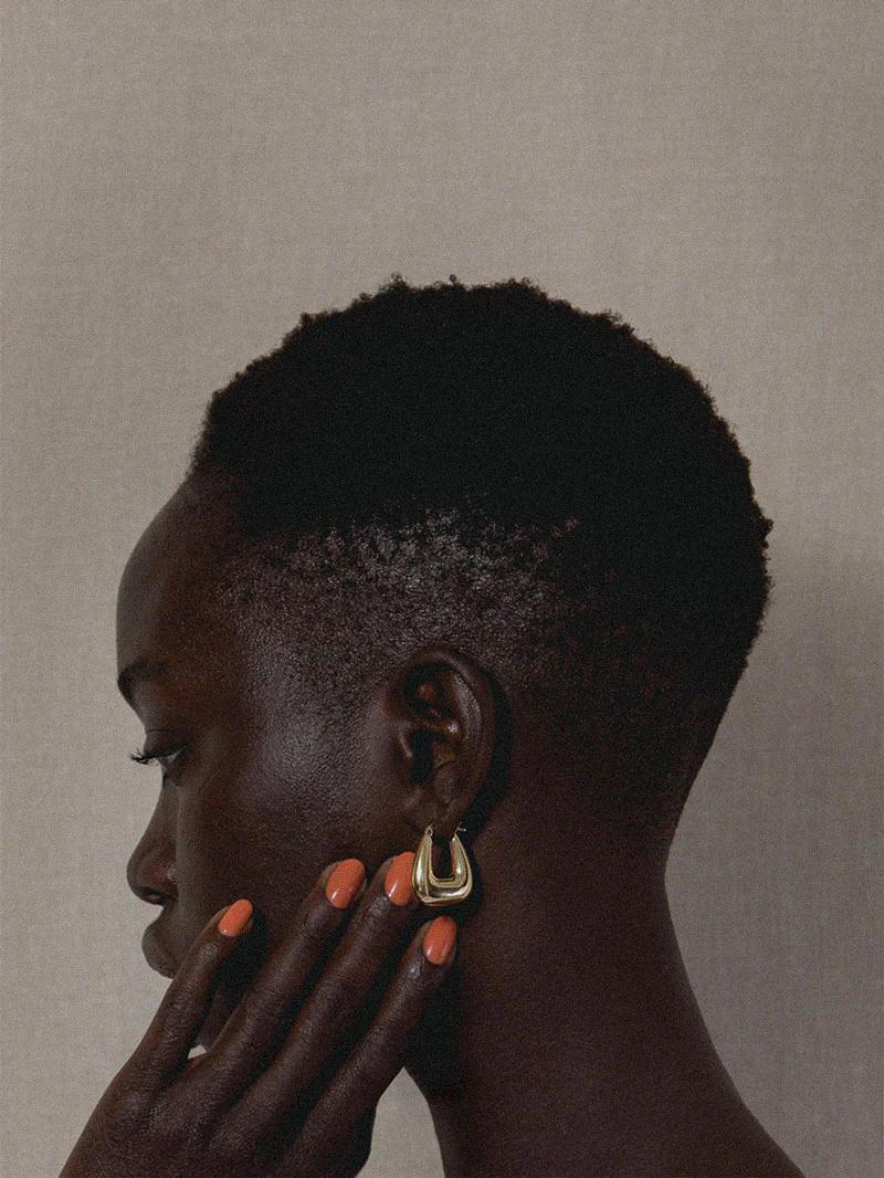 Profile view of a person with short hair wearing a gold horseshoe earring and bright orange nails, painted with high-quality polish from J. Hannah, against a neutral background.