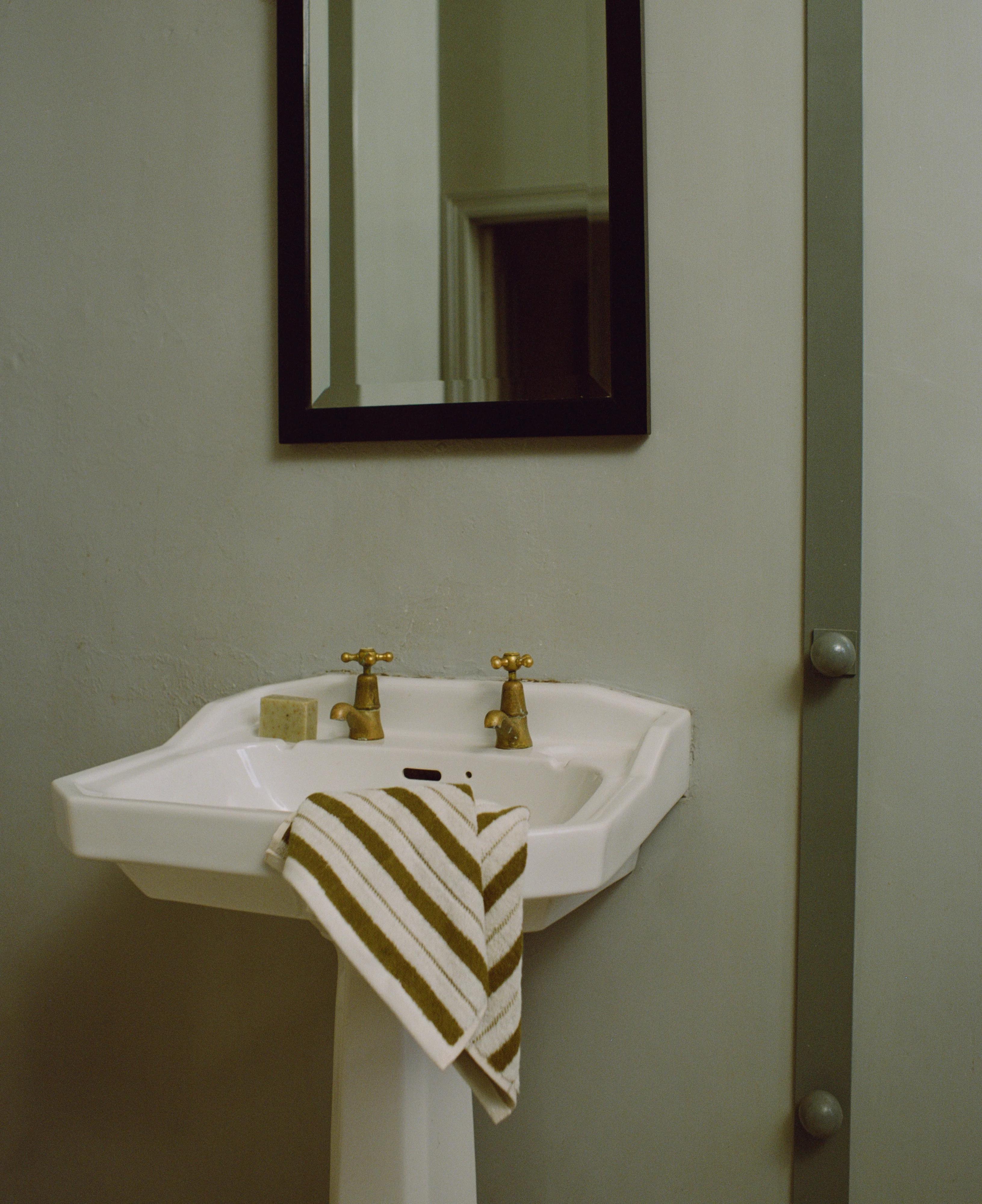 White pedestal sink with dual brass faucets. Rectangular mirror above. Striped towel draped over edge.