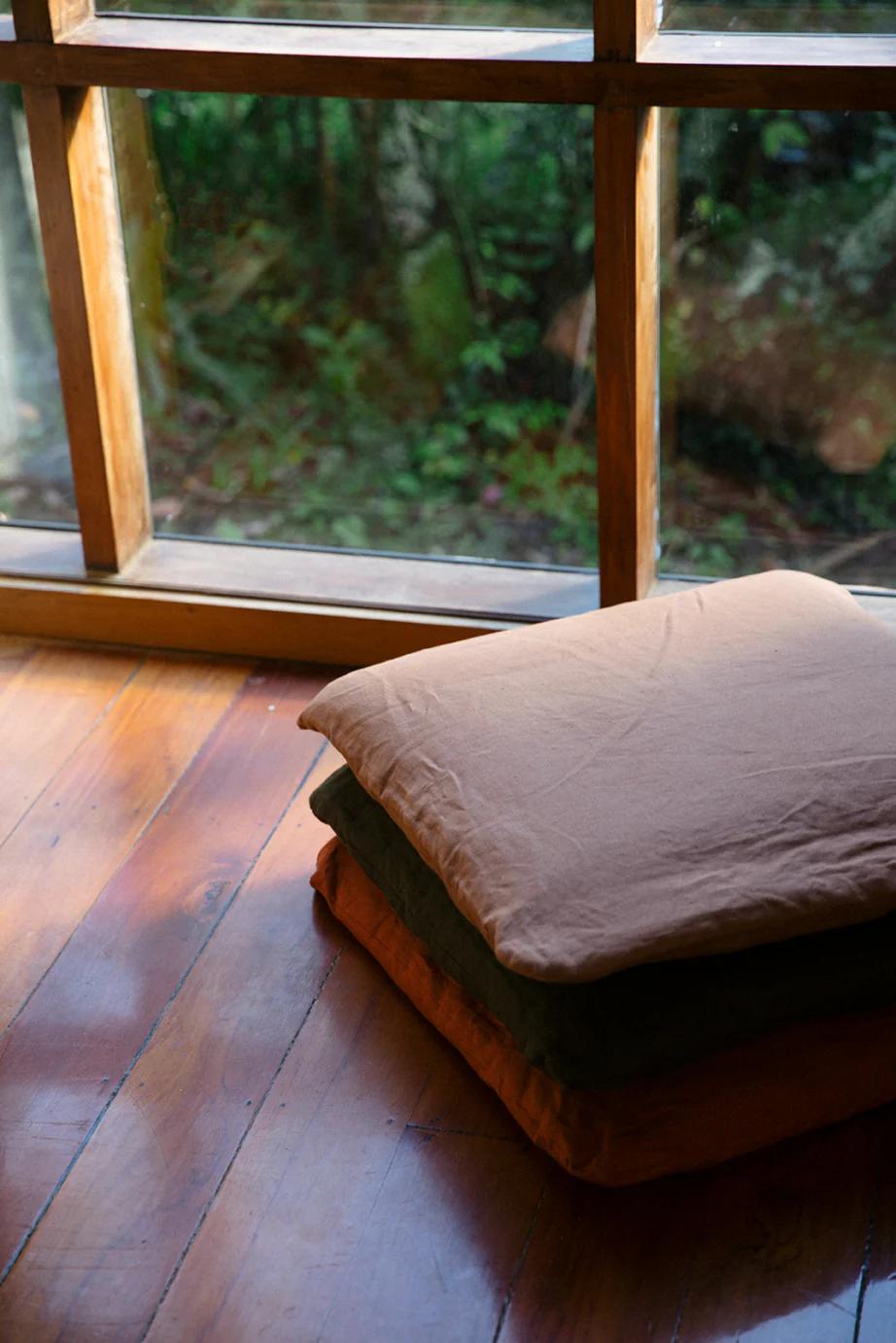 Three KLAY cushions in earthy tones stacked on wooden floor. Large window frames view of lush garden, blending indoor coziness with nature.