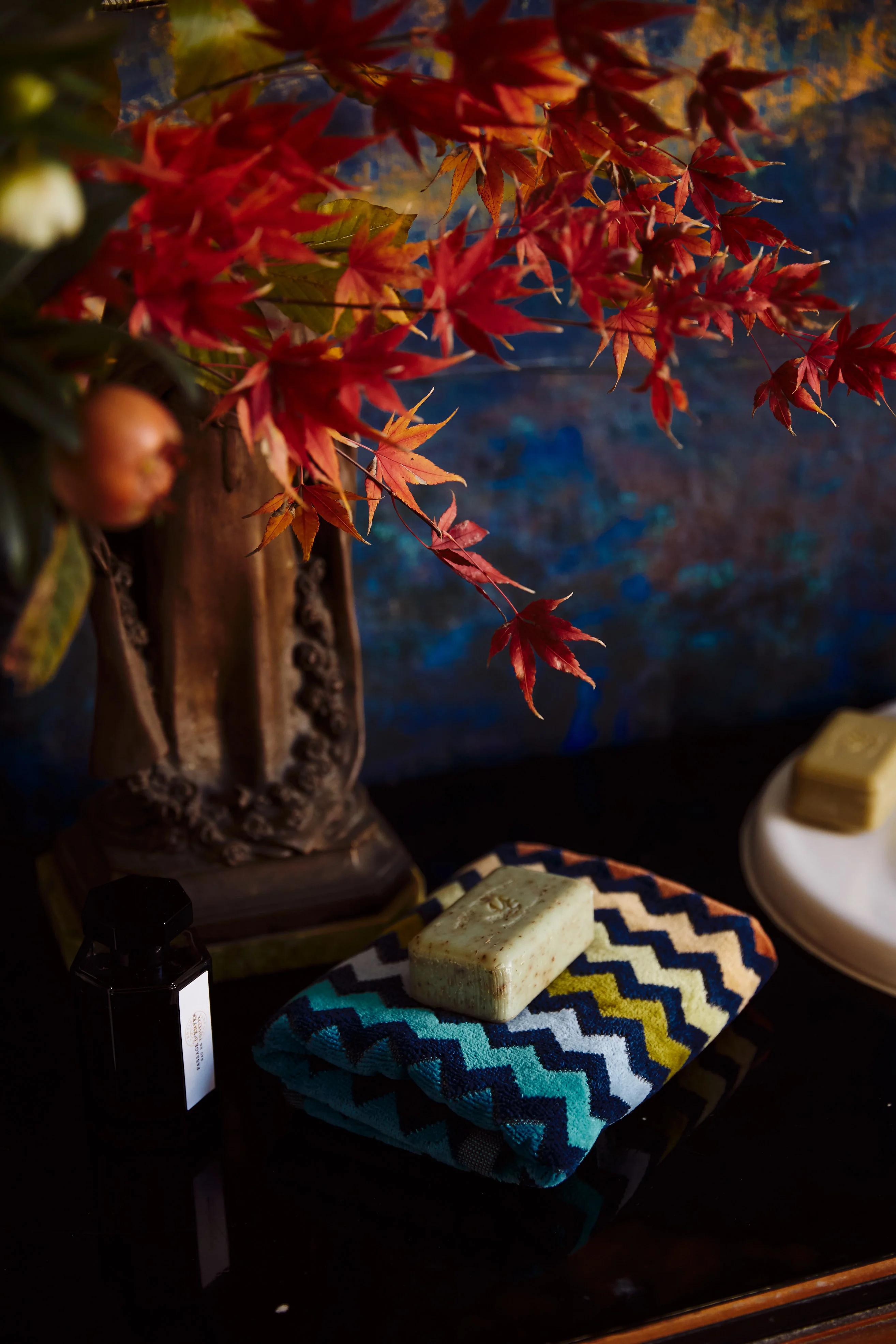 Dark scene with zigzag towel, soap, and vase of autumn leaves. Textured blue wall background evokes deep winter solstice hues.