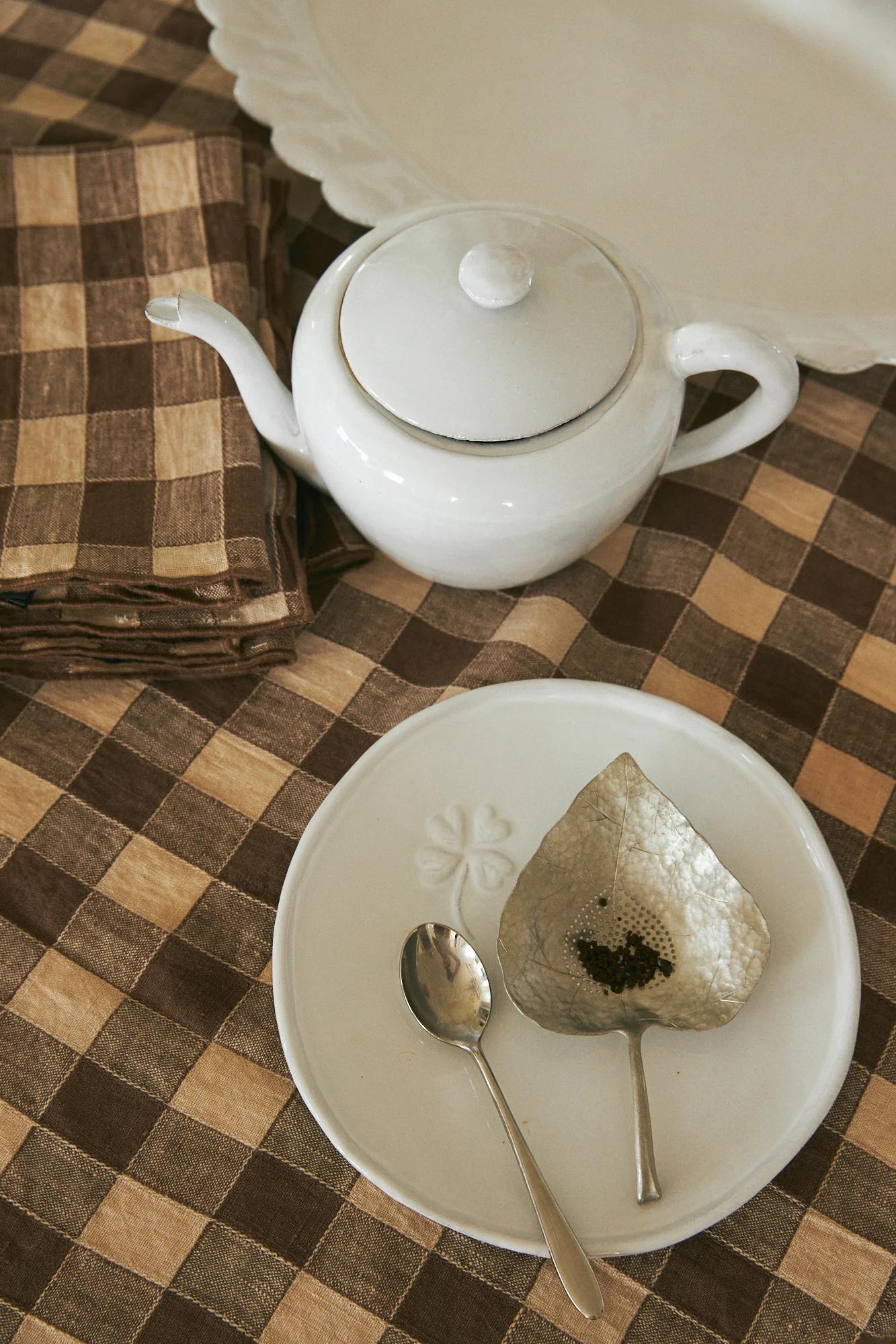 White teapot beside stacked checkered napkins. White plate with spoons and pepper. Checkered tablecloth.