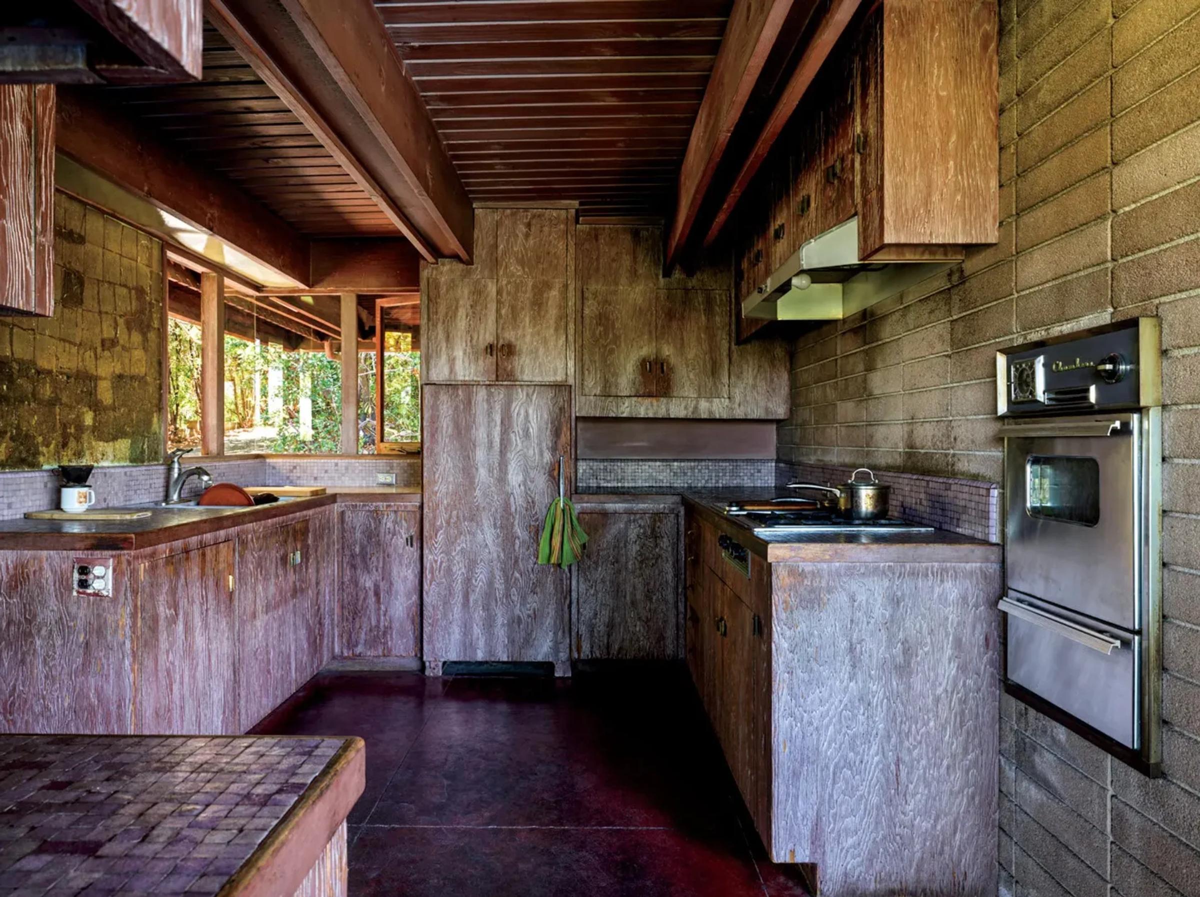 A rustic kitchen with entirely wooden cabinetry and brick walls. Features an embedded oven stove, refrigerator, and sink. Sunlight filters through large windows to the left.