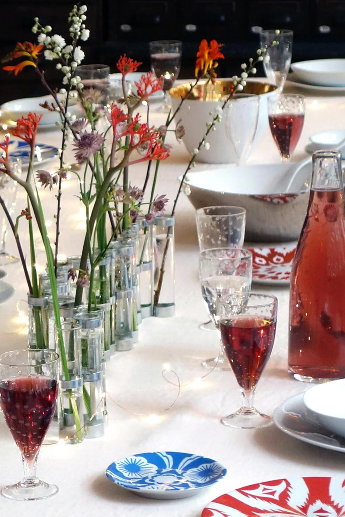 Elegant table setting with red and white flowers, glasses of red drinks, and patterned plates. Fairy lights add ambiance.