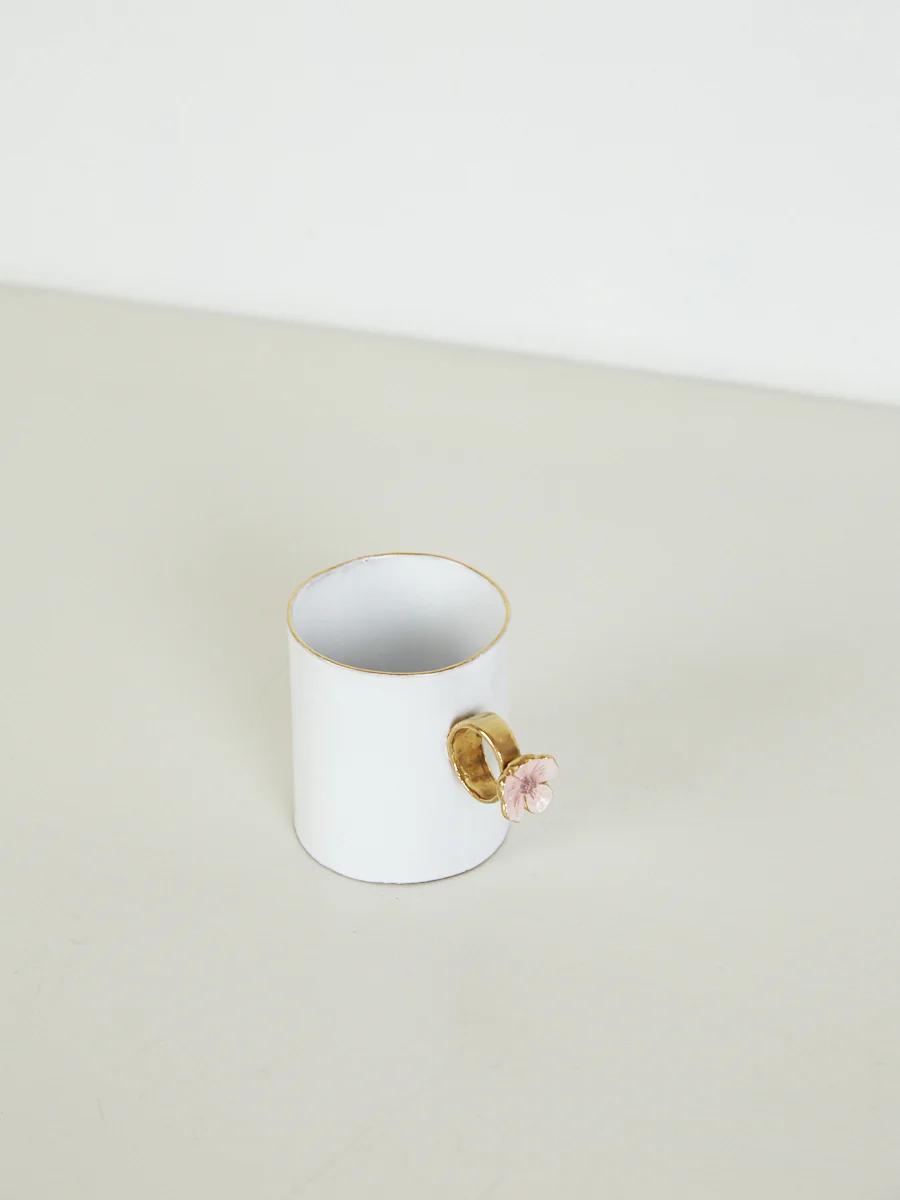 White mug with gold-lined handle and pink flower decor on light surface. Plain, light background.