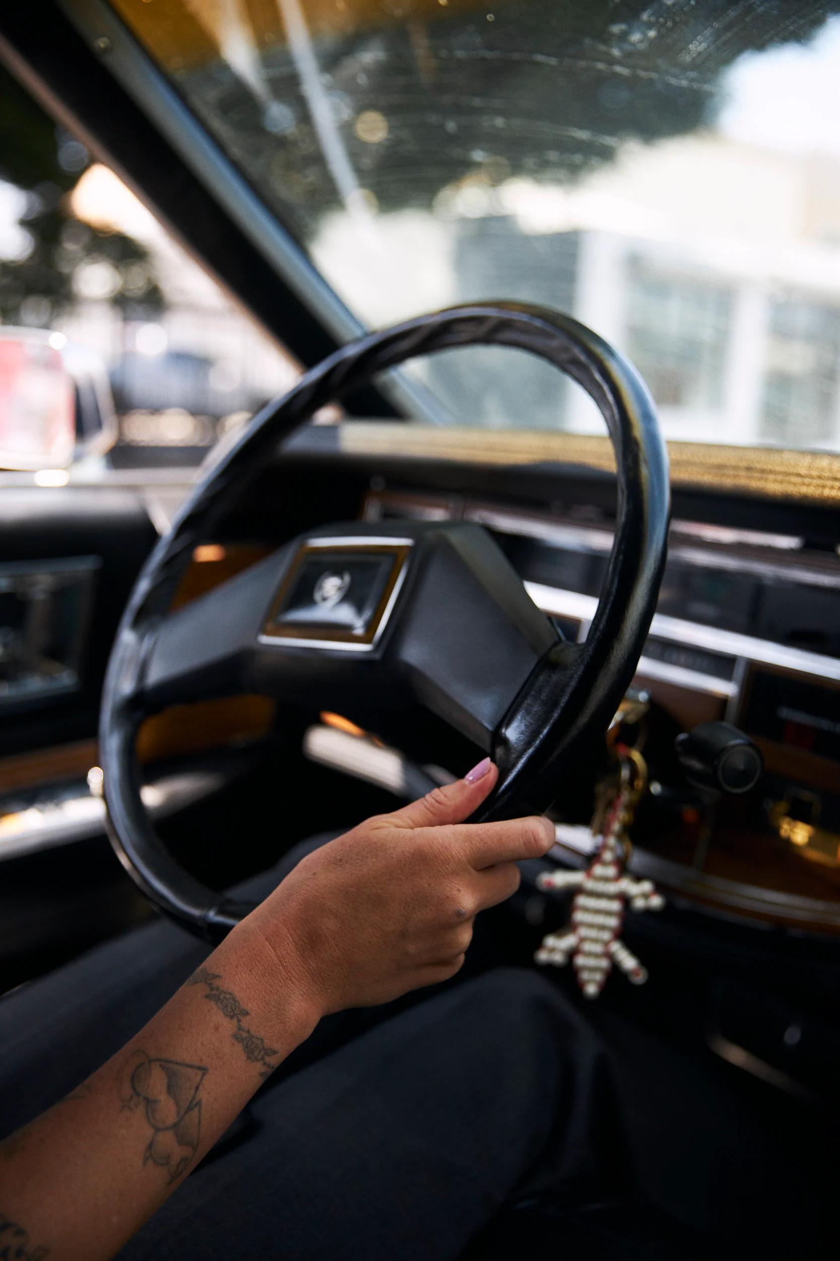 Tattooed arm holds classic car steering wheel. Striped decoration hangs from ignition. Wooden trim on dashboard.