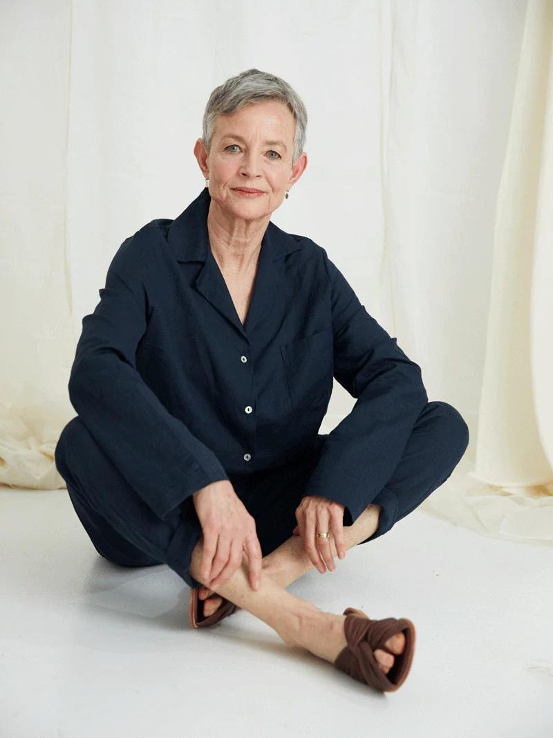 Mature woman with short gray hair sits cross-legged on floor. Dark blue outfit, brown sandals. Calm expression, light background.