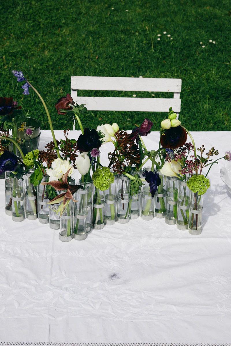 White chair behind long table with white cloth. Small vases of wild flowers, green grass background.